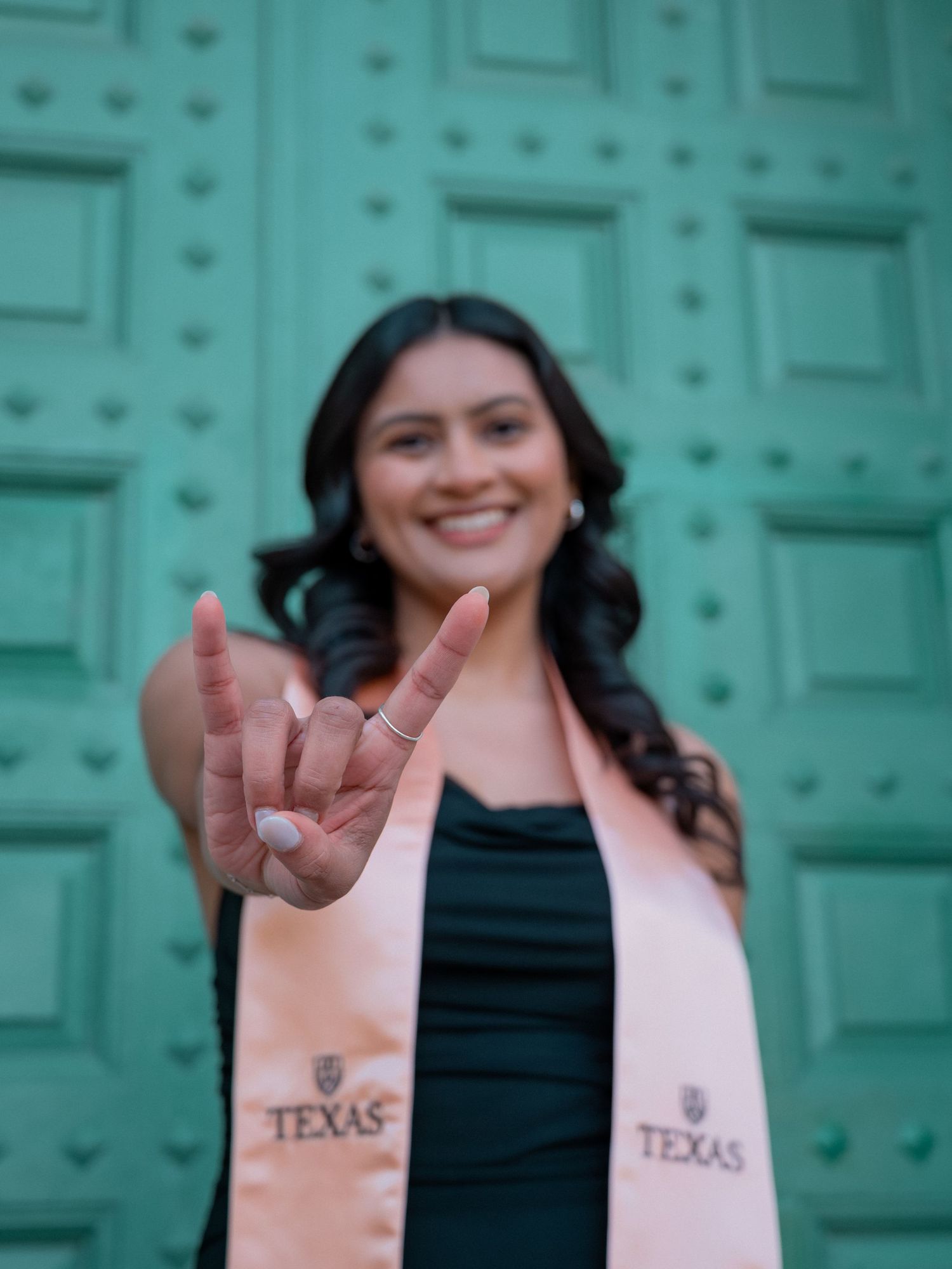 Graduate wearing a Texas graduation stole makes a rock and roll hand gesture in front of a turquoise door.