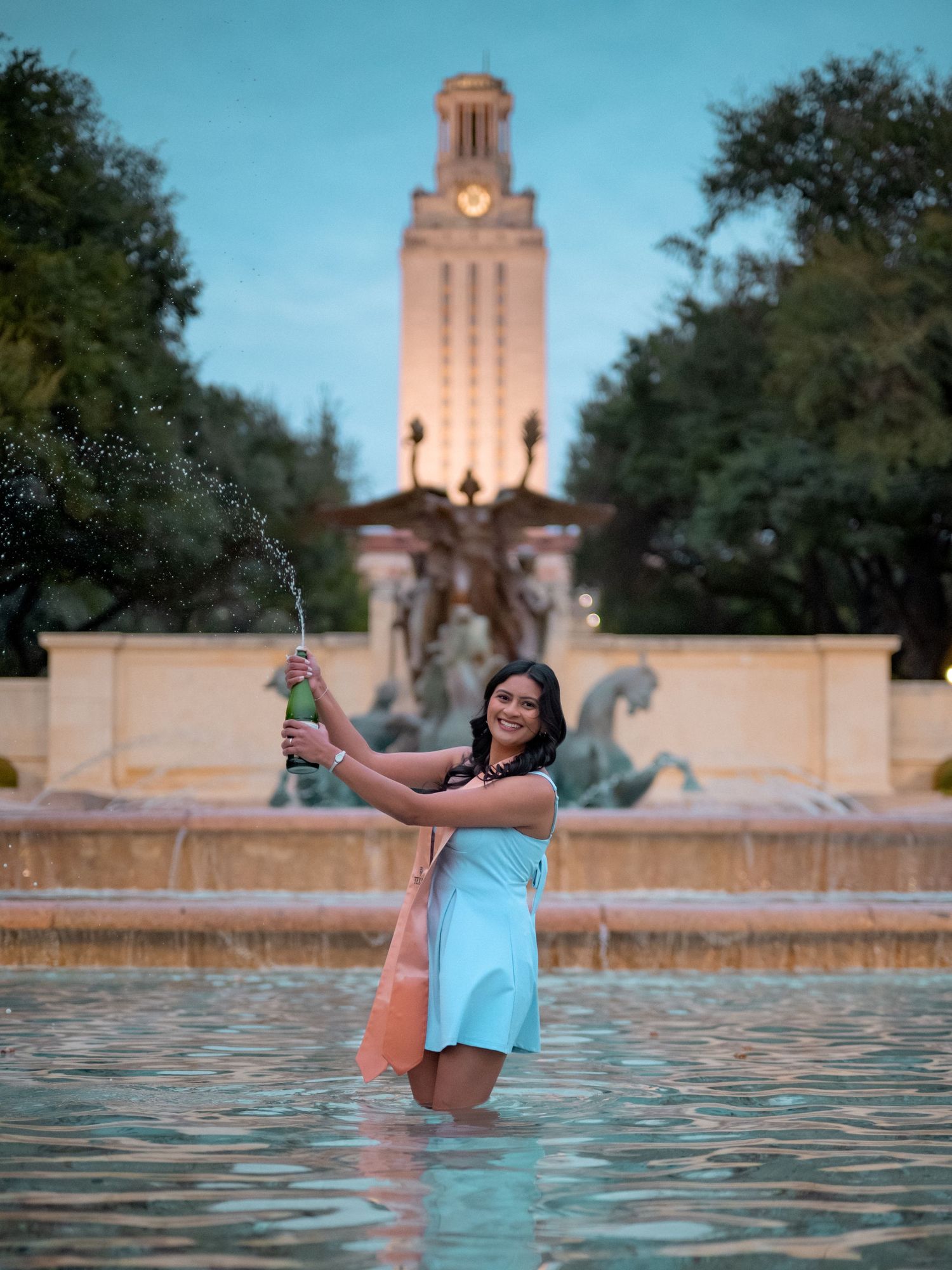 A person enjoys a refreshing swim in the UT Austin Tower fountain at sunset with the iconic tower illuminated in the background.