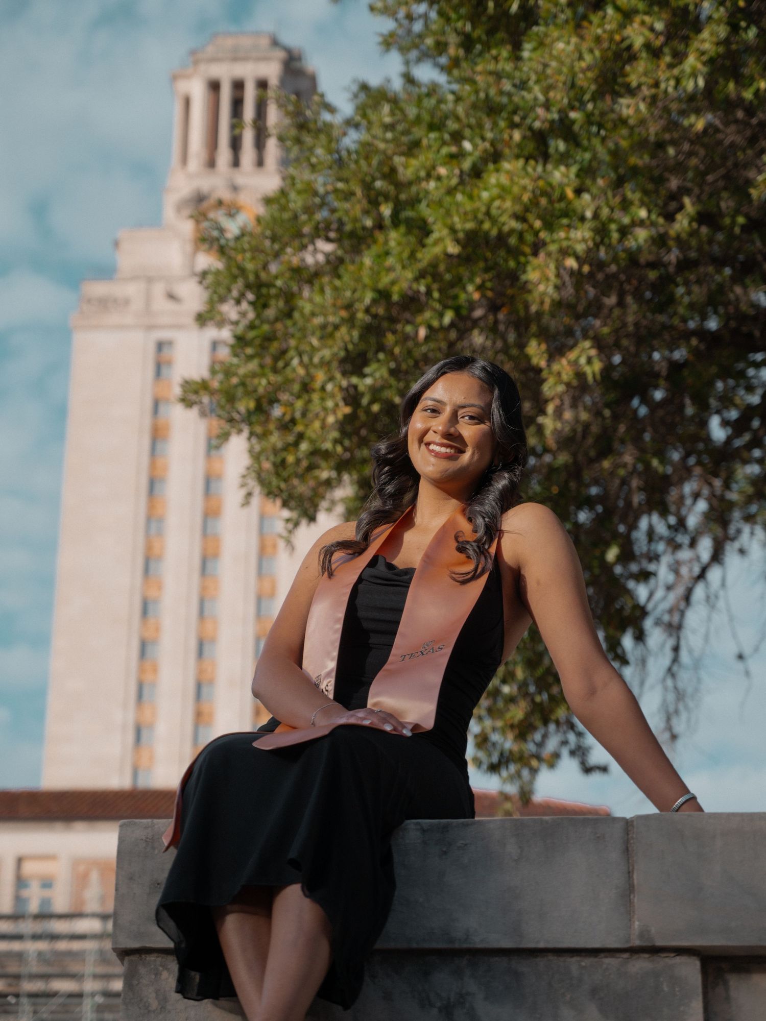 A student in a black dress and pink sash sits on a stone wall with the iconic UT Austin Tower in the background.