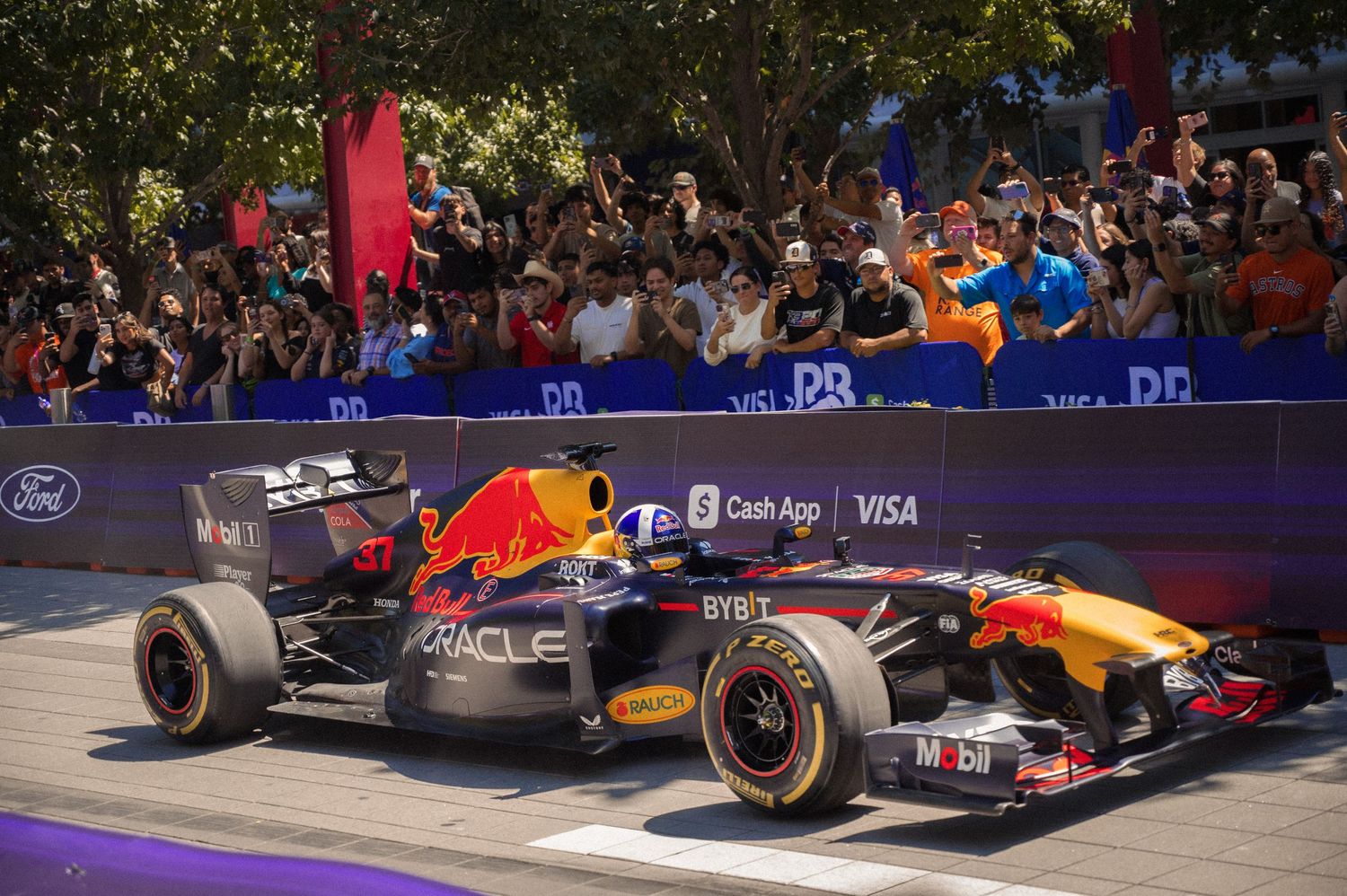 Red Bull Formula 1 car performs demonstration run on city streets with spectators watching from barriers.