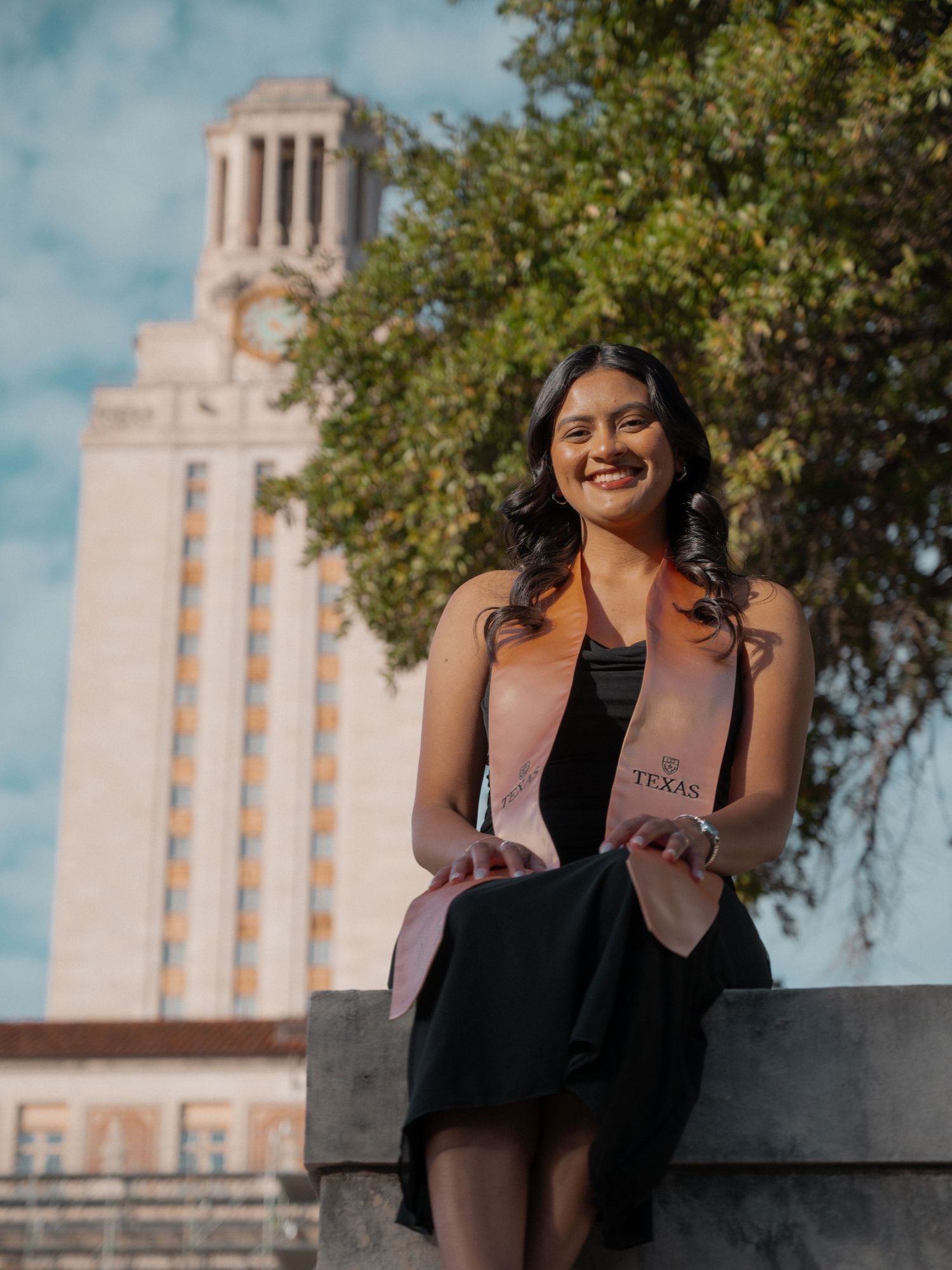A graduate stands smiling in front of the iconic University of Texas Tower on a sunny day.