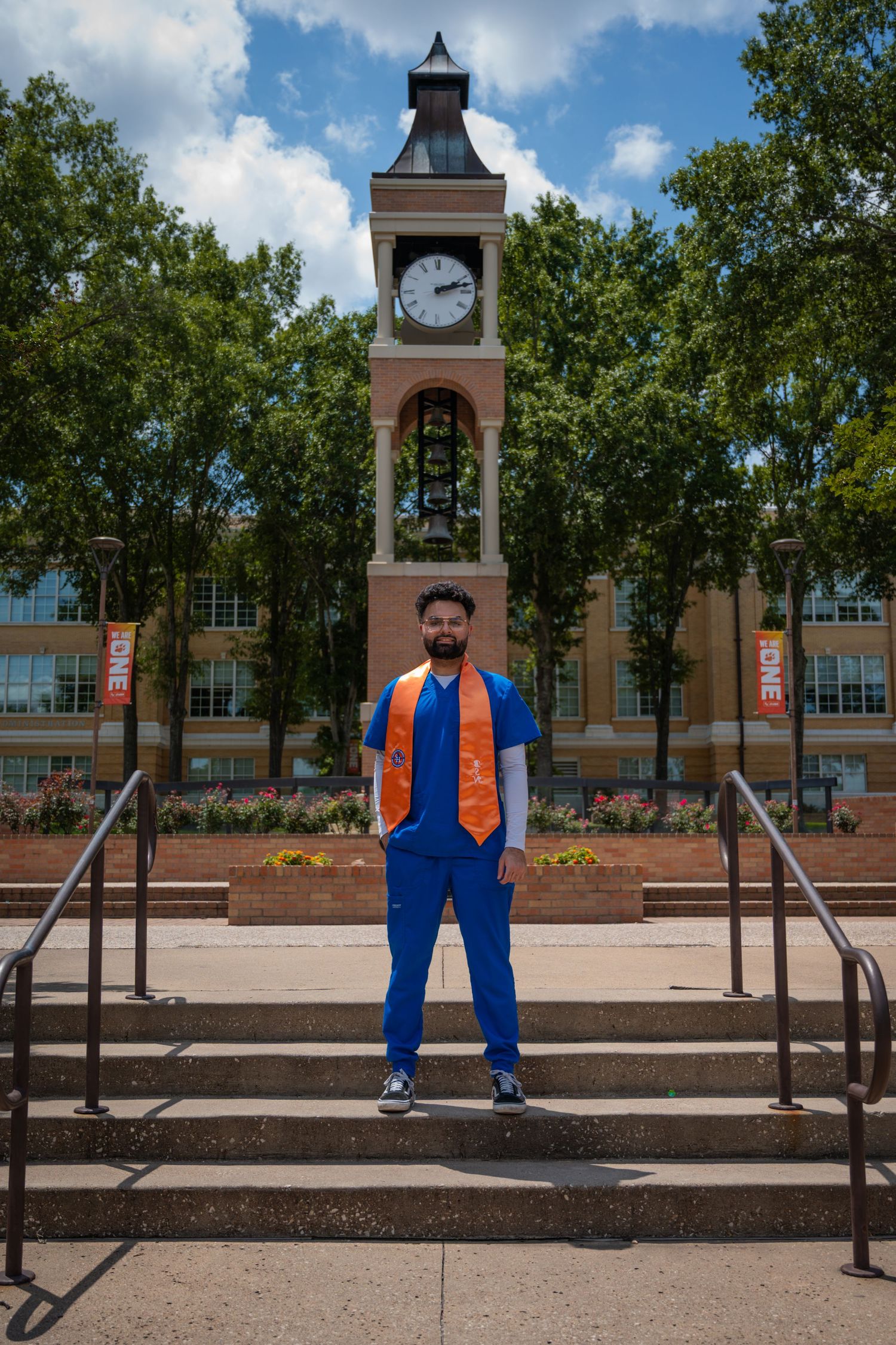 Healthcare professional in blue scrubs and orange stole stands on steps in front of campus clock tower.