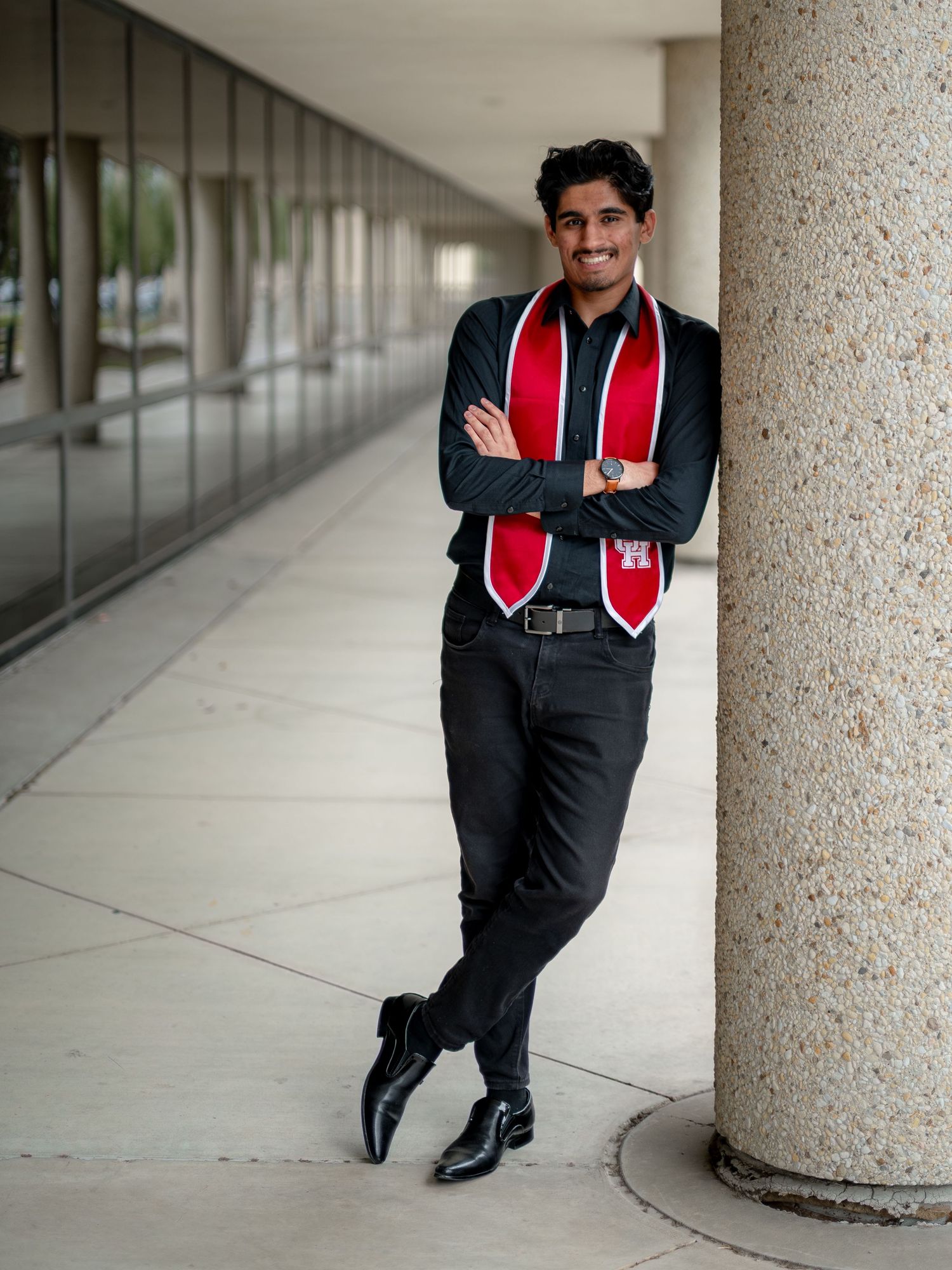 Graduate in black gown and red stole posing playfully along a columned corridor.