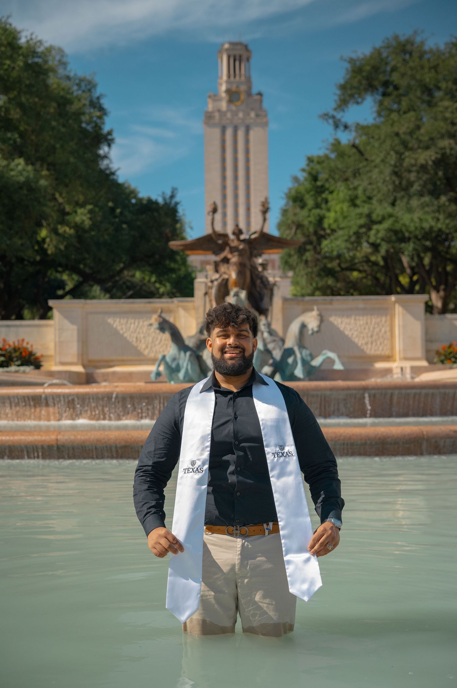 Graduate stands in fountain wearing graduation stole with UT Tower and statues behind.