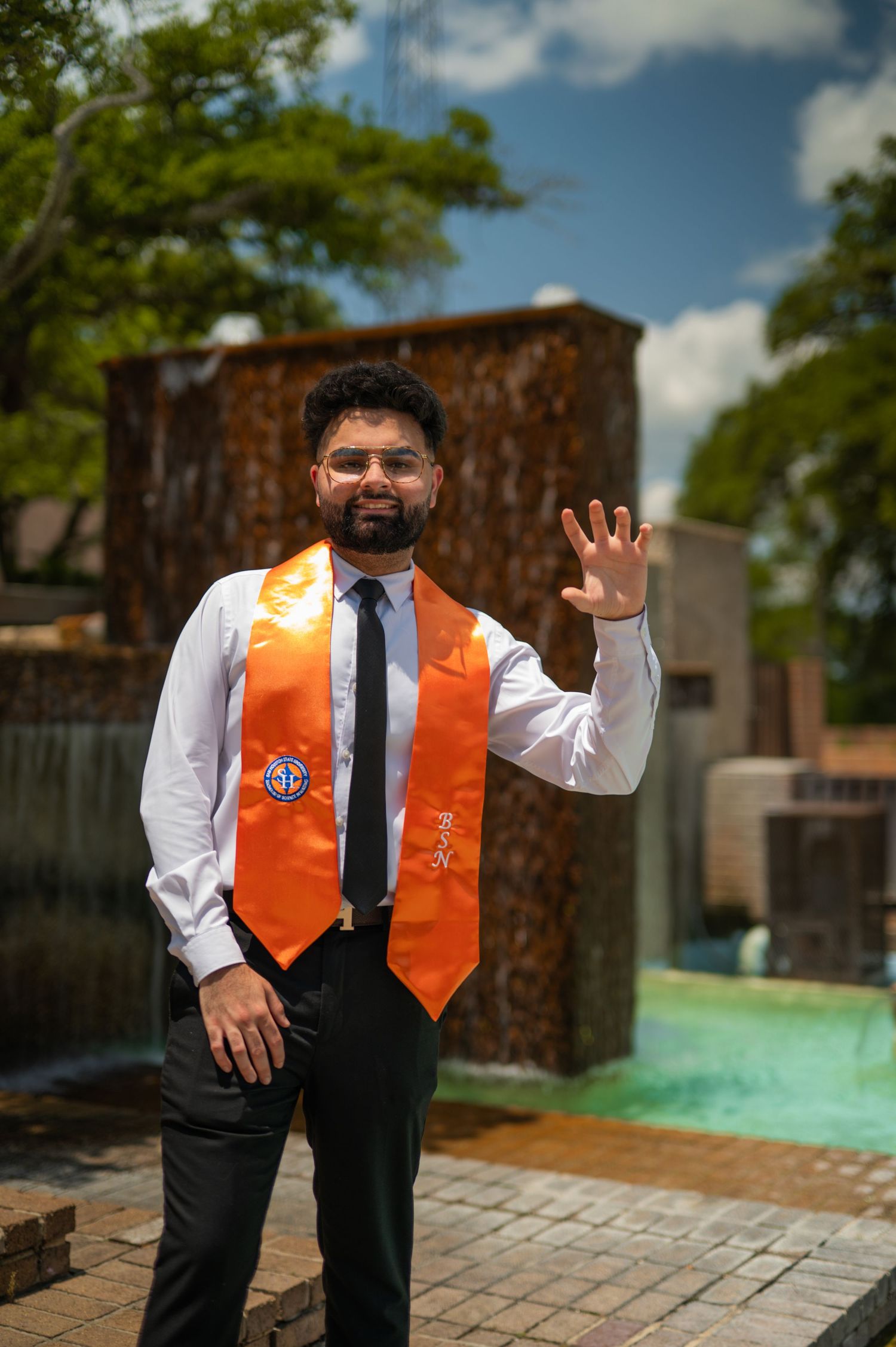 Graduate in formal wear and orange stole poses expressively outdoors on campus.