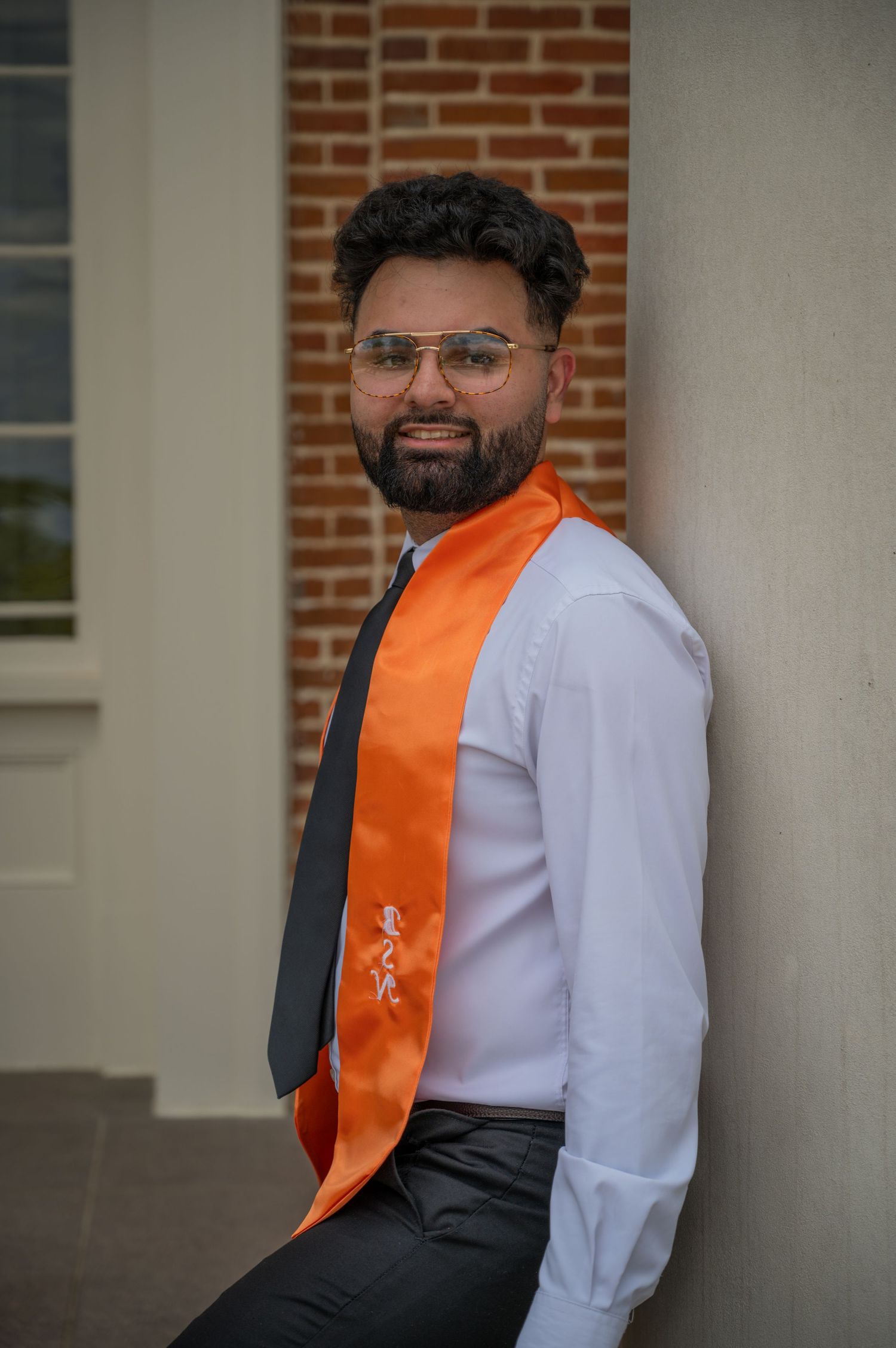Graduate wearing orange stole and white shirt smiles in front of collegiate architecture.