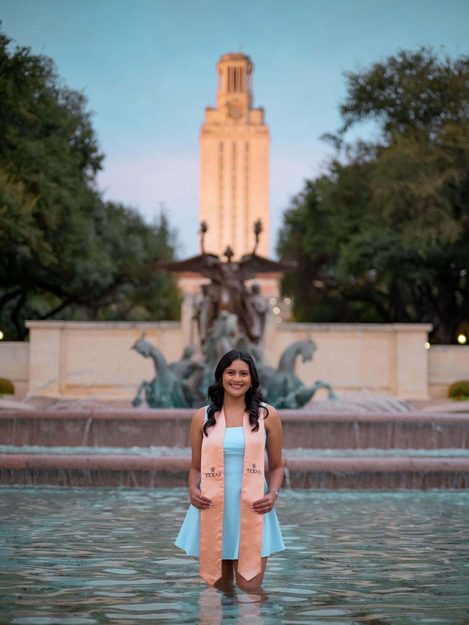 A fountain with bronze horse sculptures stands in front of the iconic UT Austin Tower at sunset.