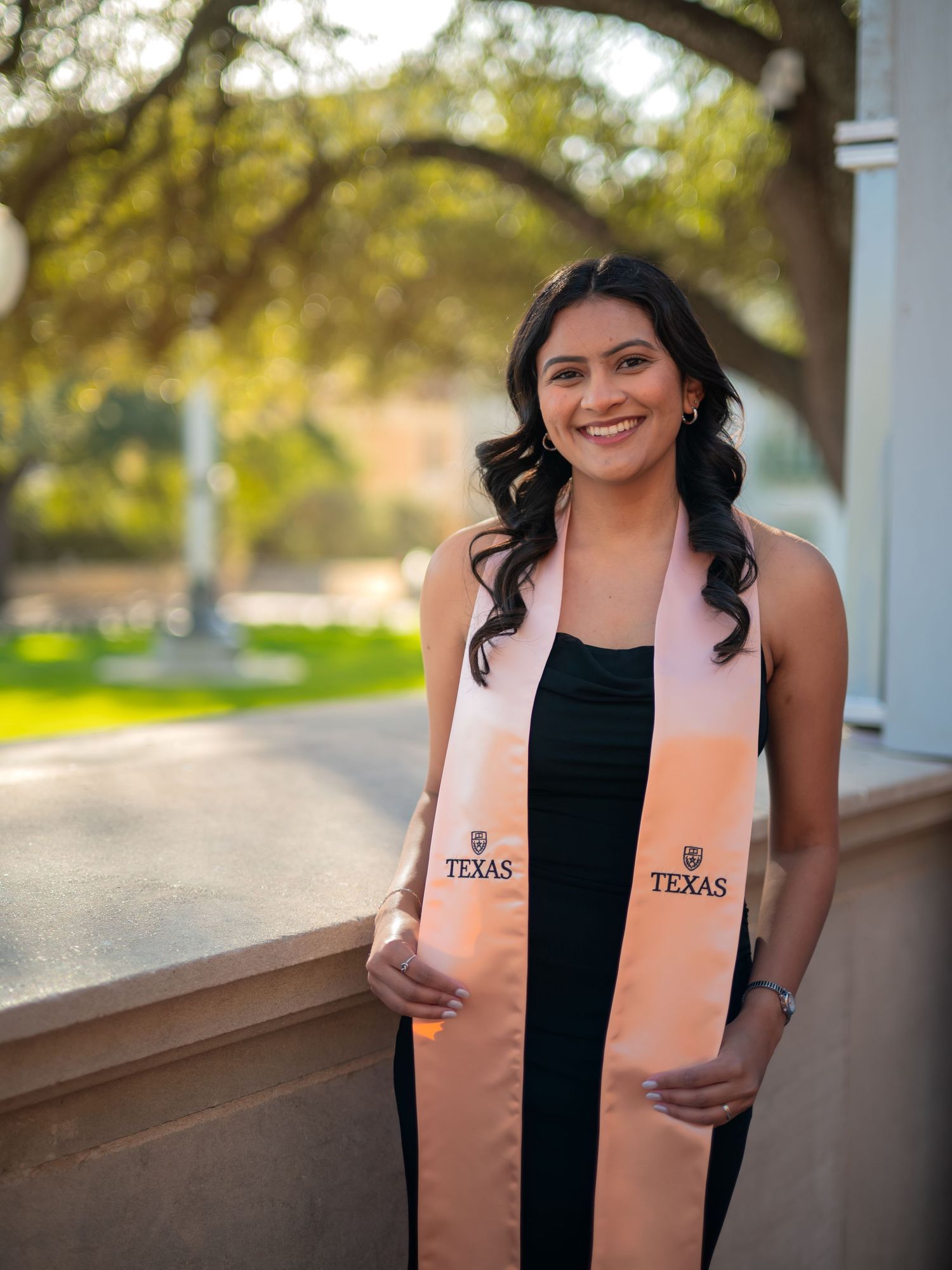 Graduate wearing a black dress and peach-colored University of Texas graduation stole poses outdoors on sunny day.