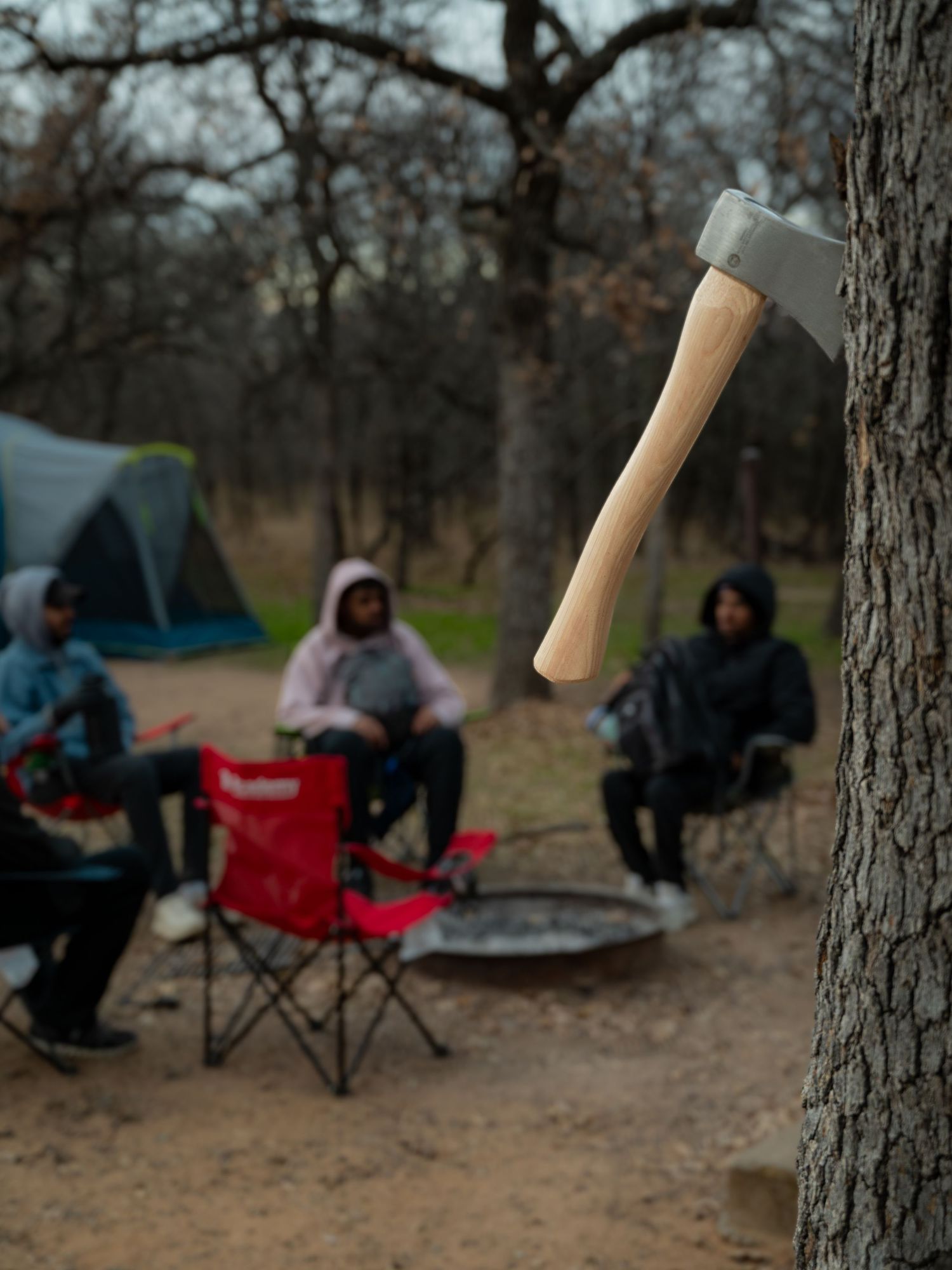 People sitting in camping chairs in a wooded area with camping equipment visible.