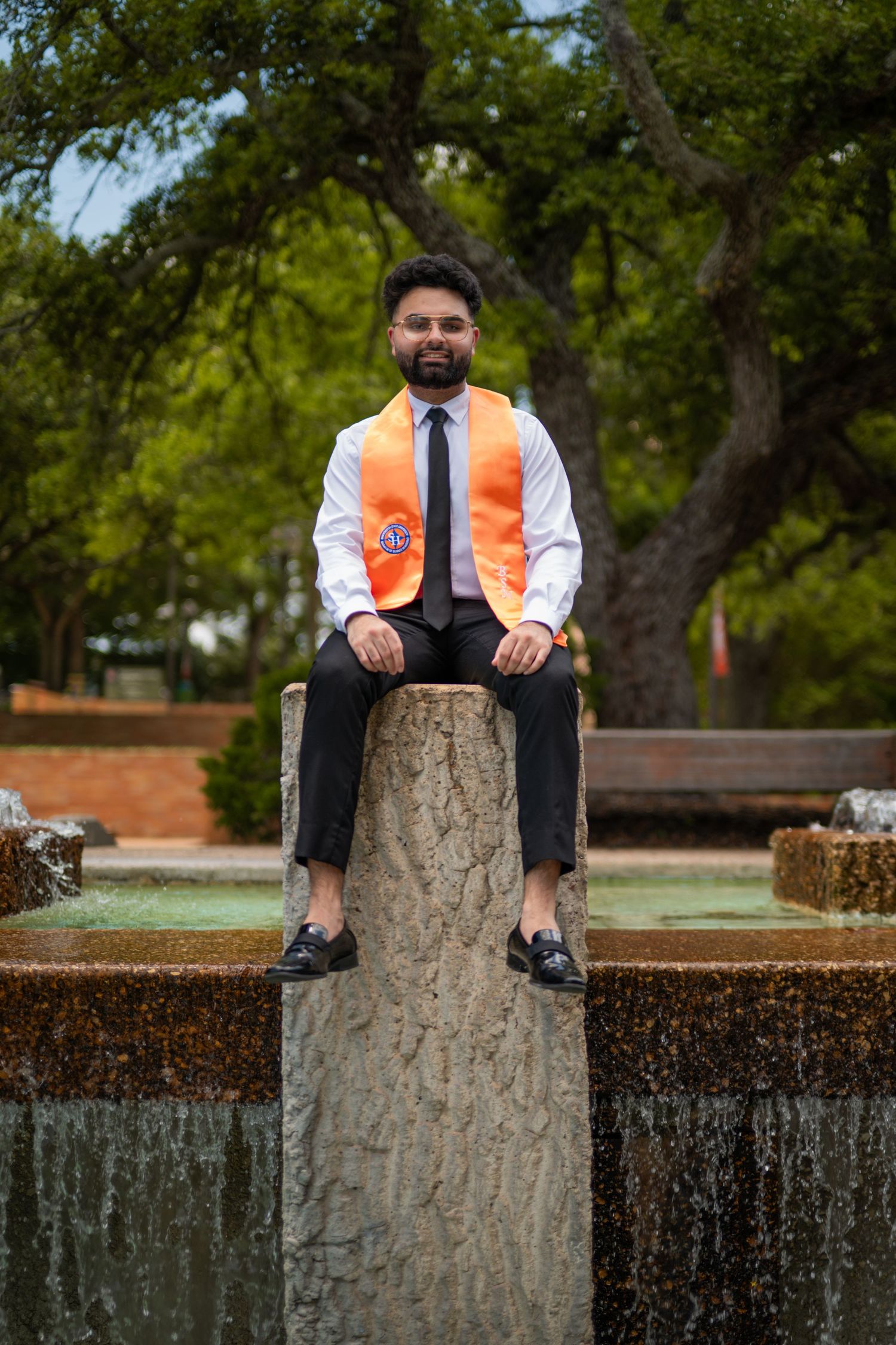 Graduate in academic regalia sits atop stone wall during outdoor photo session.