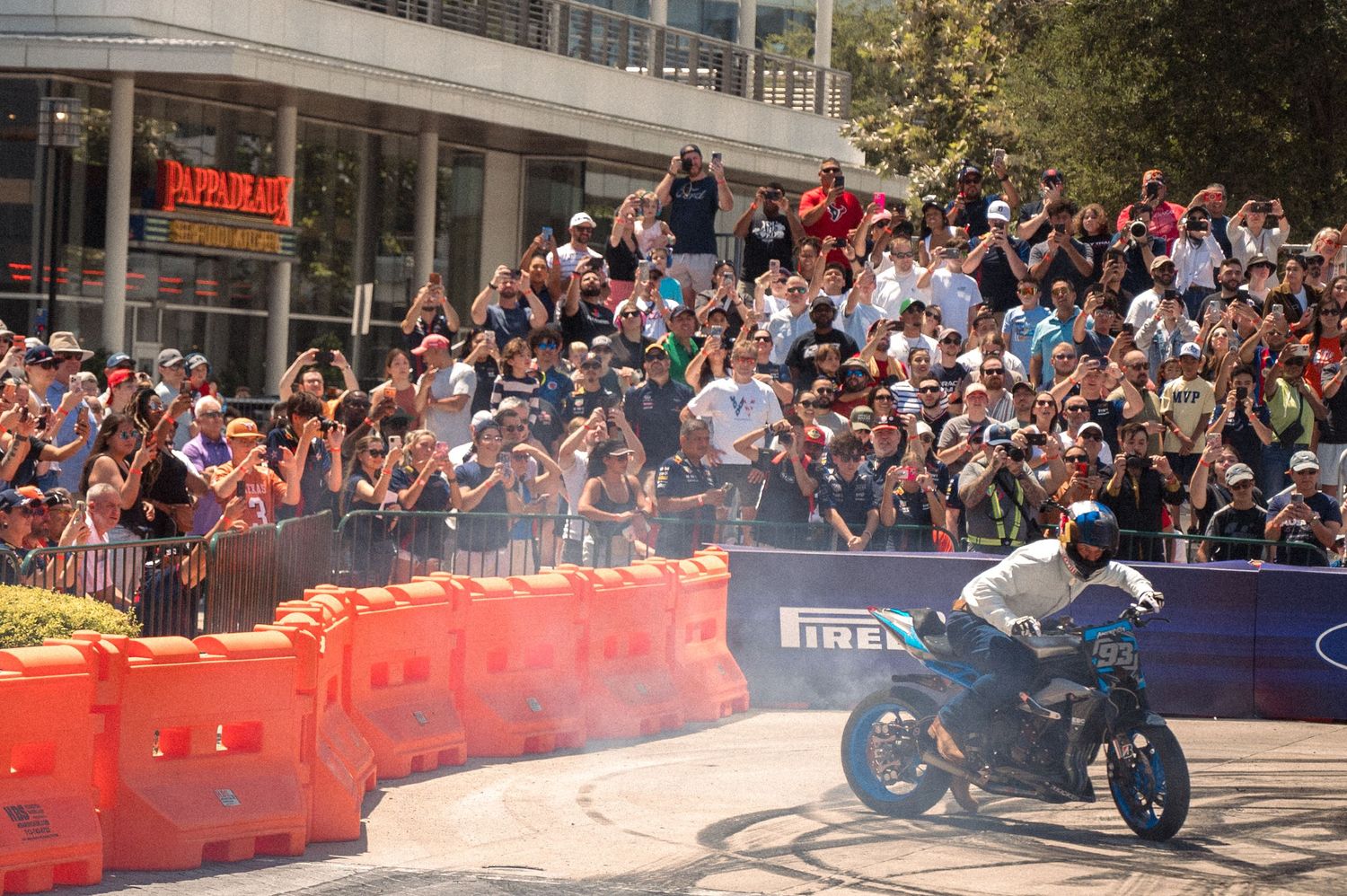 Motorcycle performing a demonstration on a street track with orange barriers and large crowd watching.