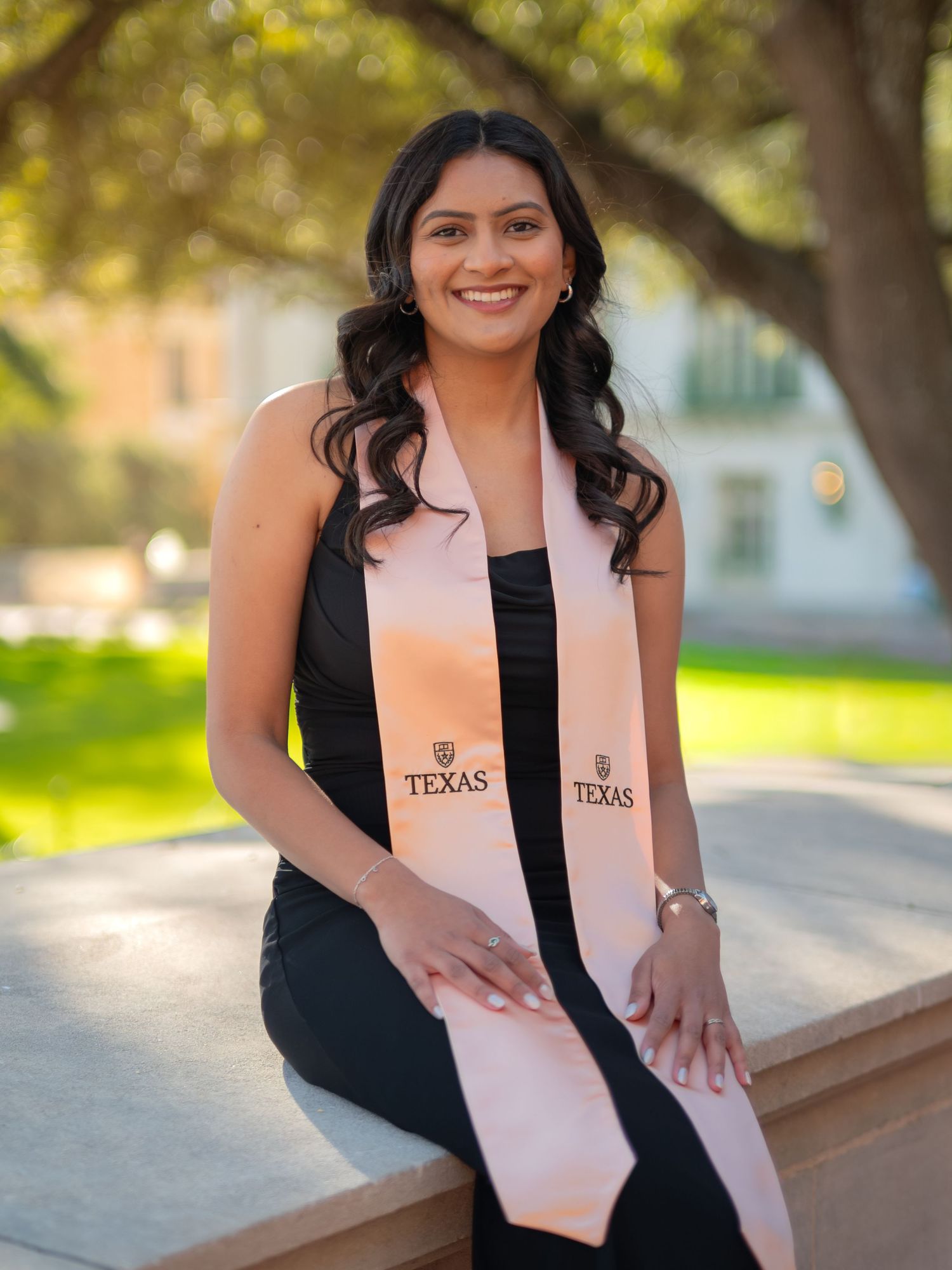 A graduate wearing a black dress and peach-colored graduation stole poses outdoors in a series of celebratory photos.