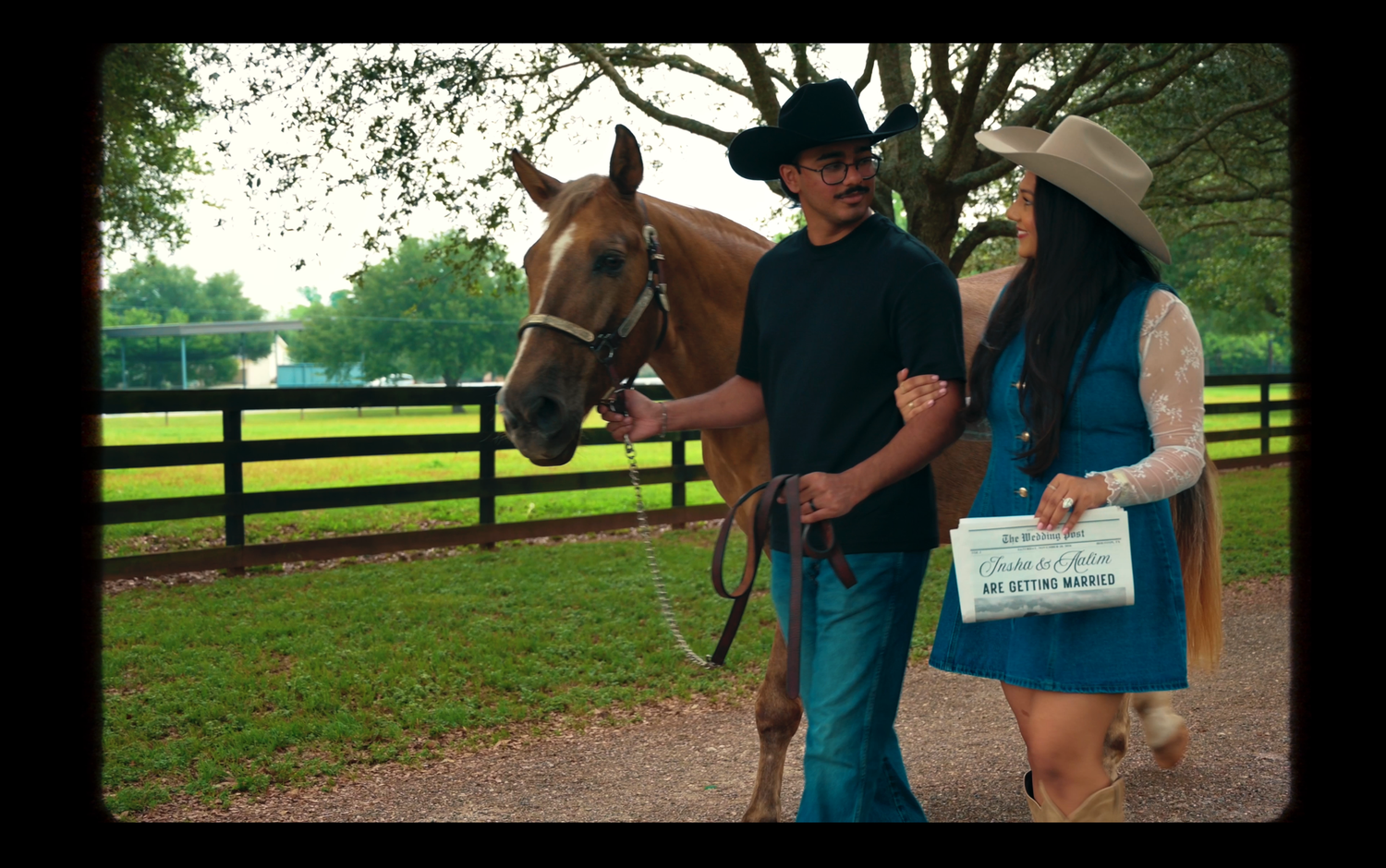 A couple in cowboy hats walks a chestnut horse along a fenced ranch path during a romantic outdoor proposal.