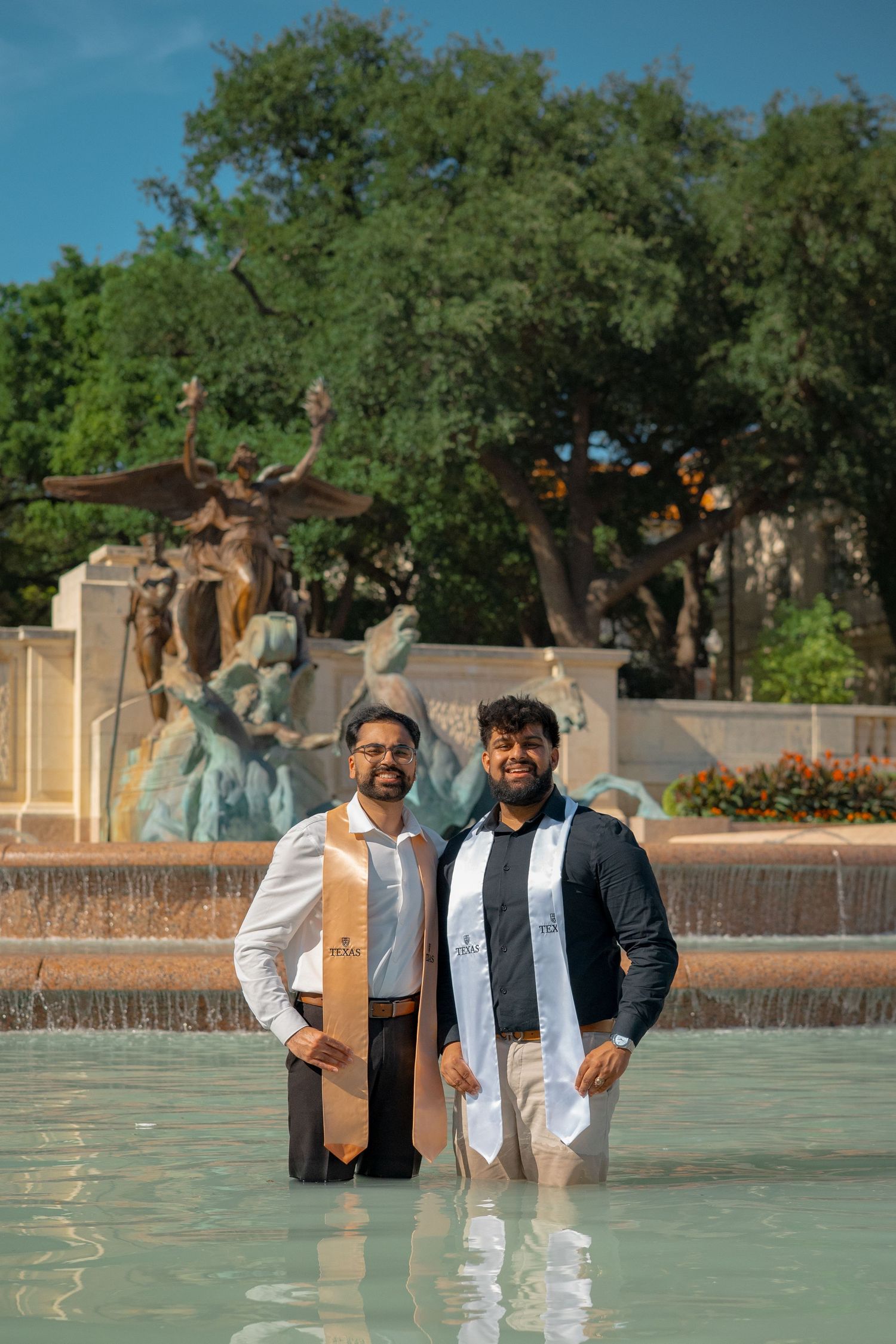 Two graduates celebrate in fountain with decorative statues and trees in background.