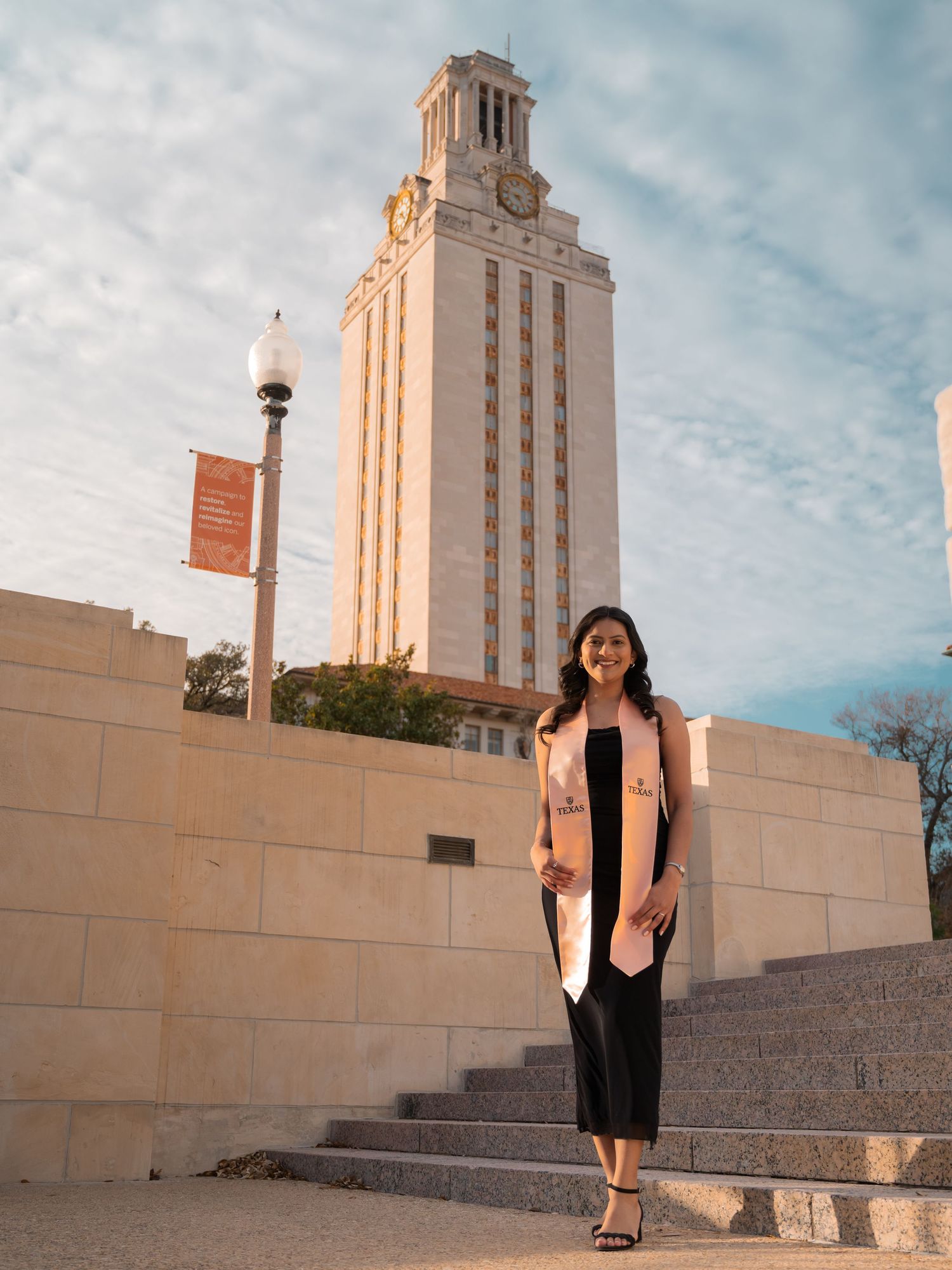 A student stands on stone steps in front of the iconic University of Texas Tower building on a sunny day.