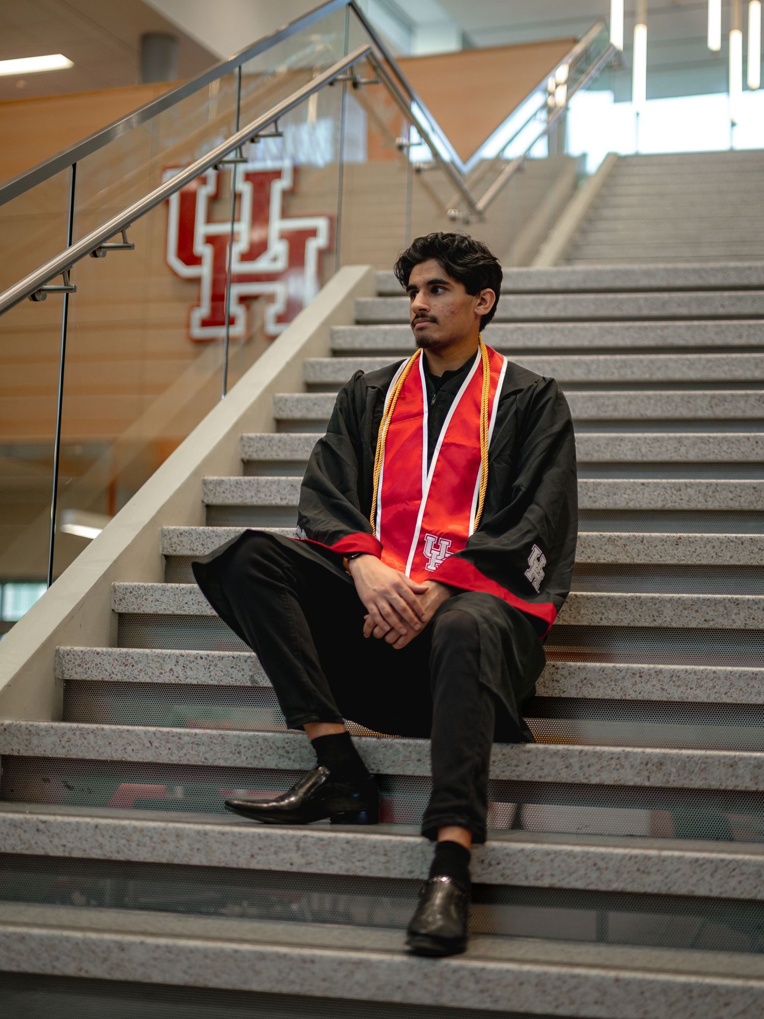 Graduate in black cap and gown with red stole sits on stairs with UH logo visible in background at University of Houston.