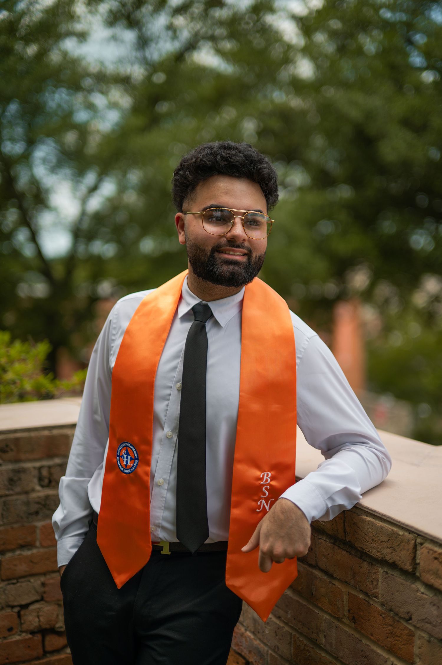Graduate in business attire with orange stole gestures while sitting on brick wall.