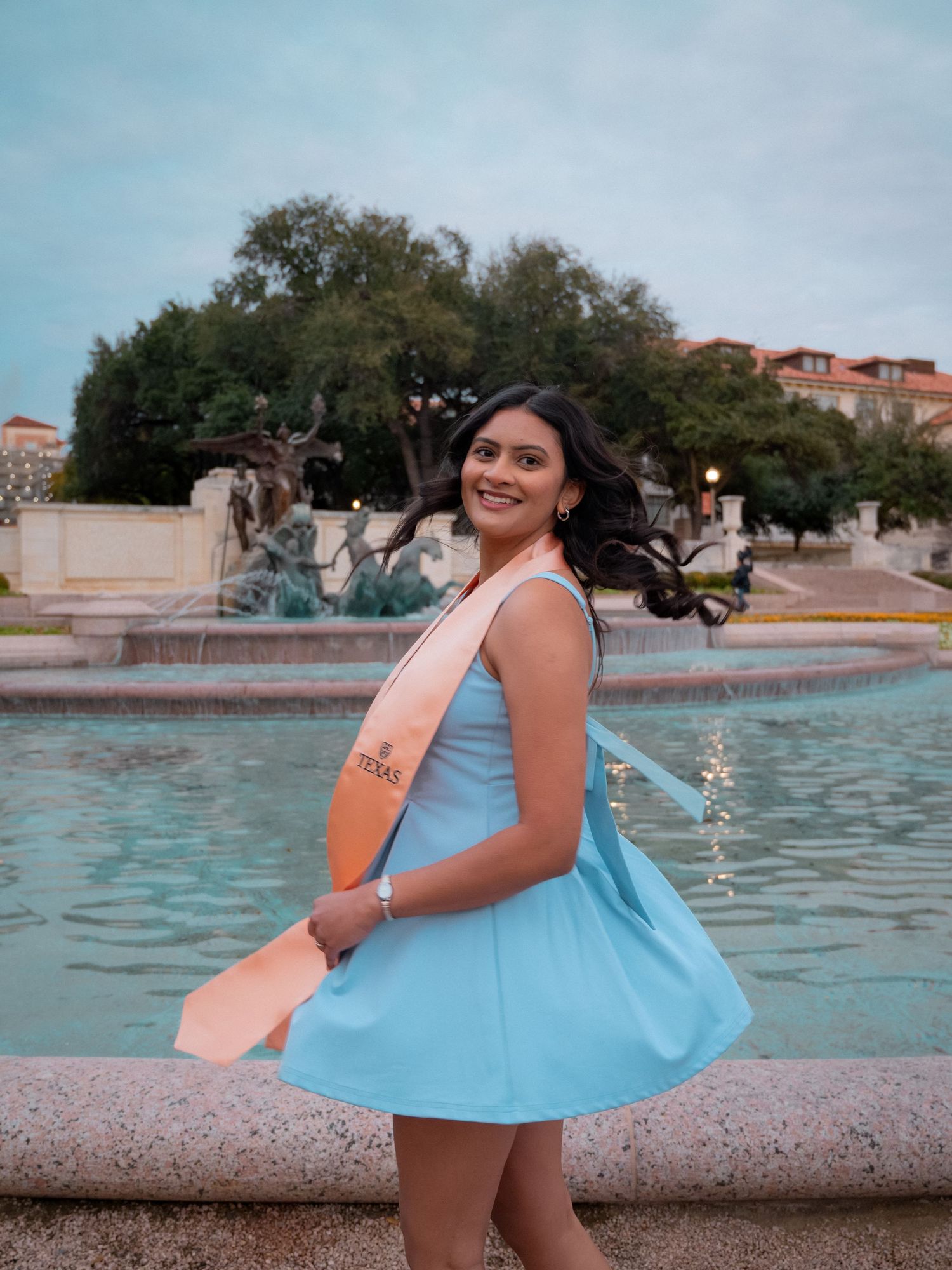 A person in a light blue dress poses playfully by a fountain on a sunny afternoon.