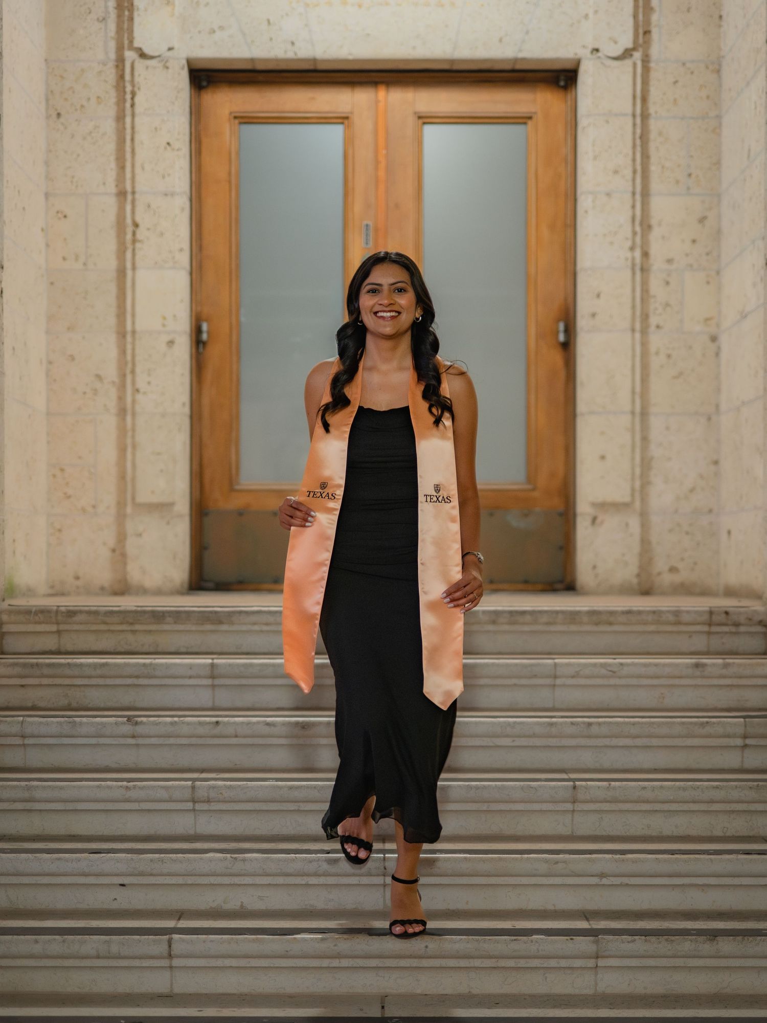 A person in a black dress and peach-colored graduation stole stands smiling on stone steps in front of wooden doors.