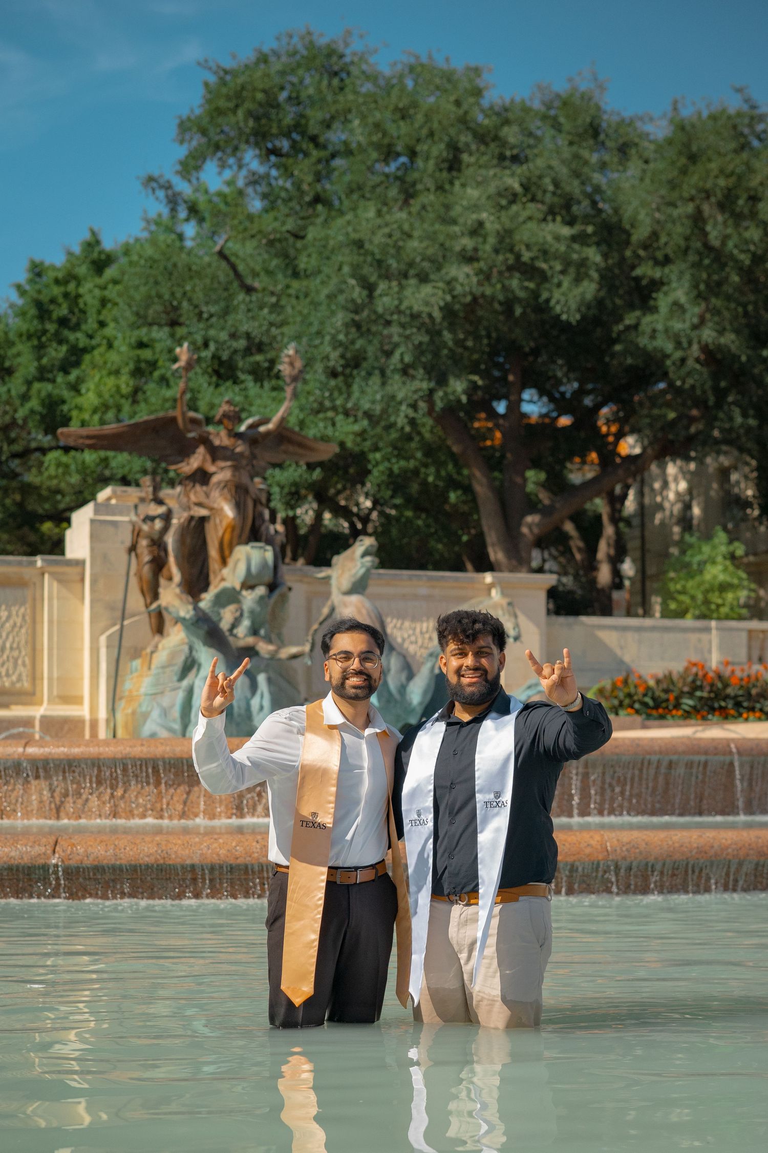 Graduates pose joyfully while standing in campus fountain with stoles and graduation attire.