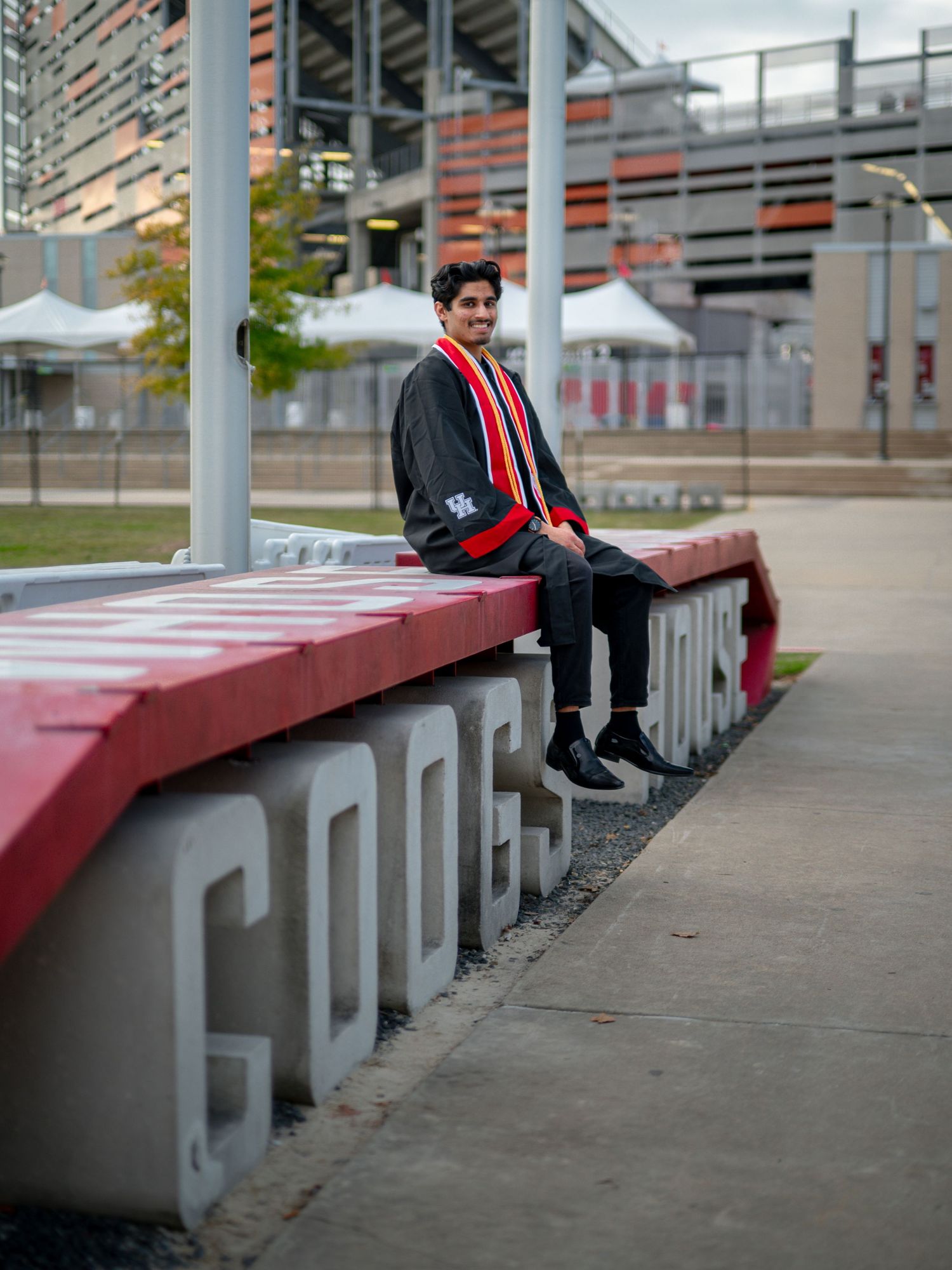 Graduate in academic regalia sitting on red concrete bench outside university building.