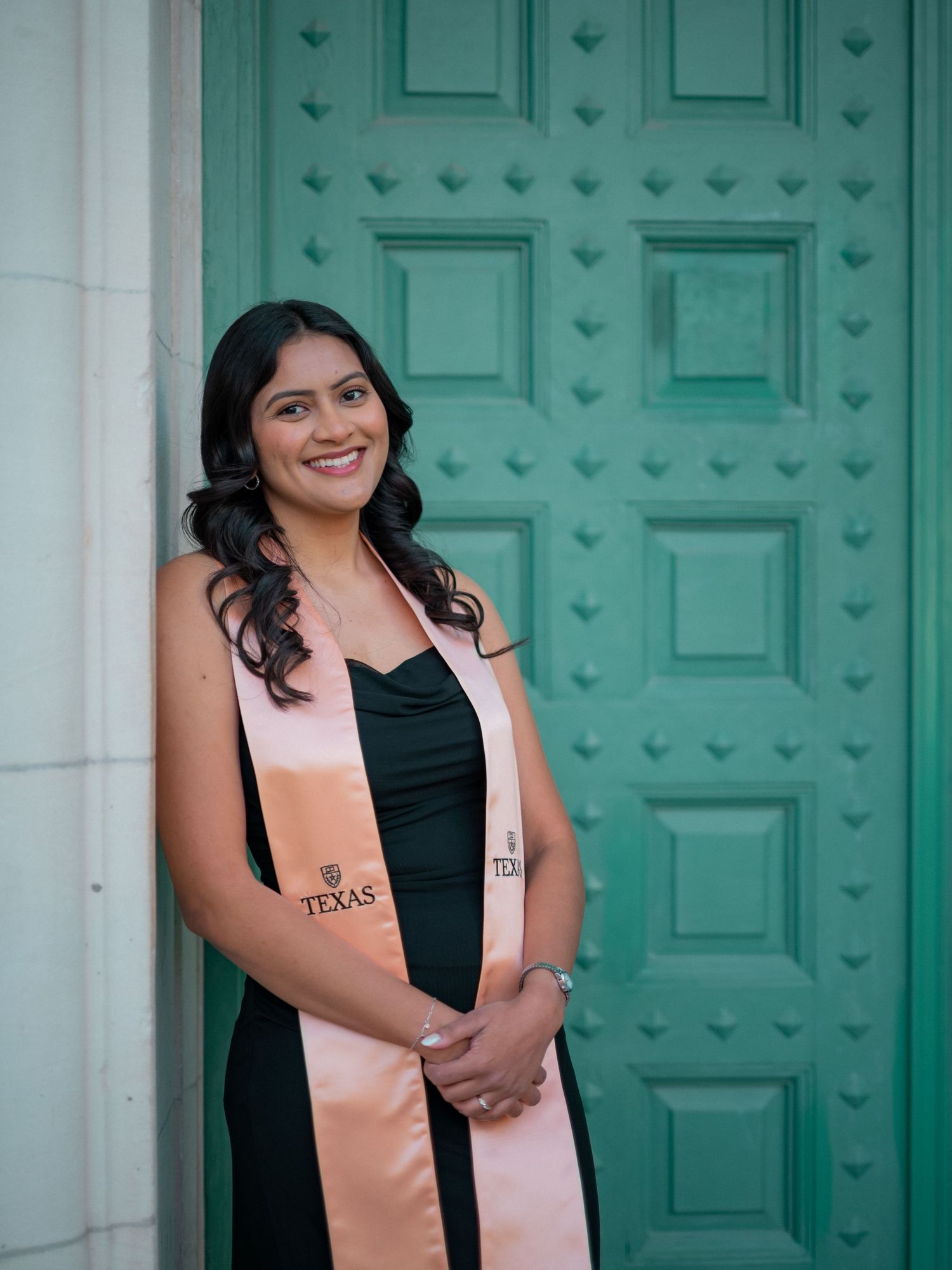 Graduate in black dress and peach stole poses for photos in front of ornate turquoise doors.