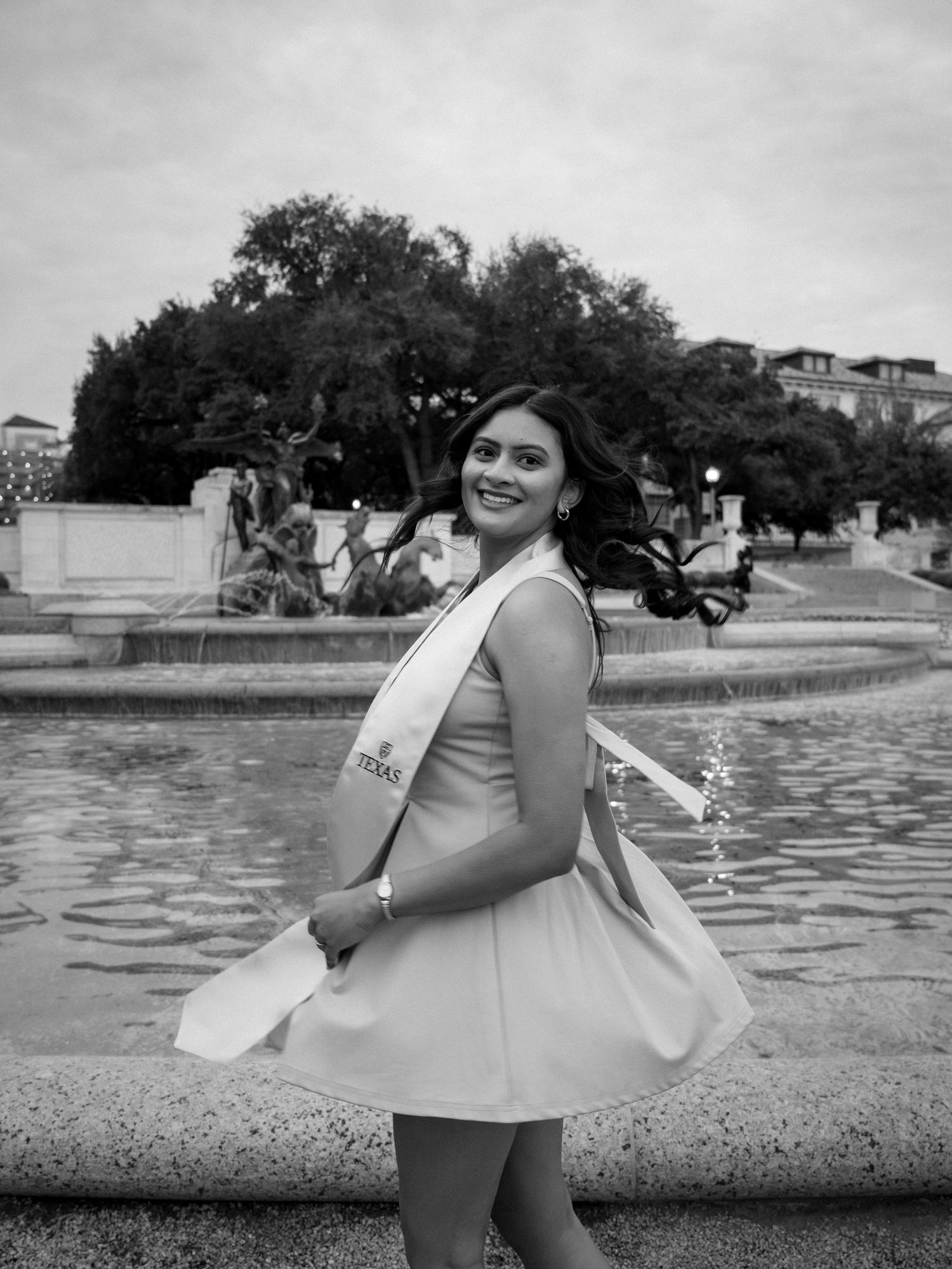 A person in a white dress poses joyfully by a fountain in a black and white photograph.