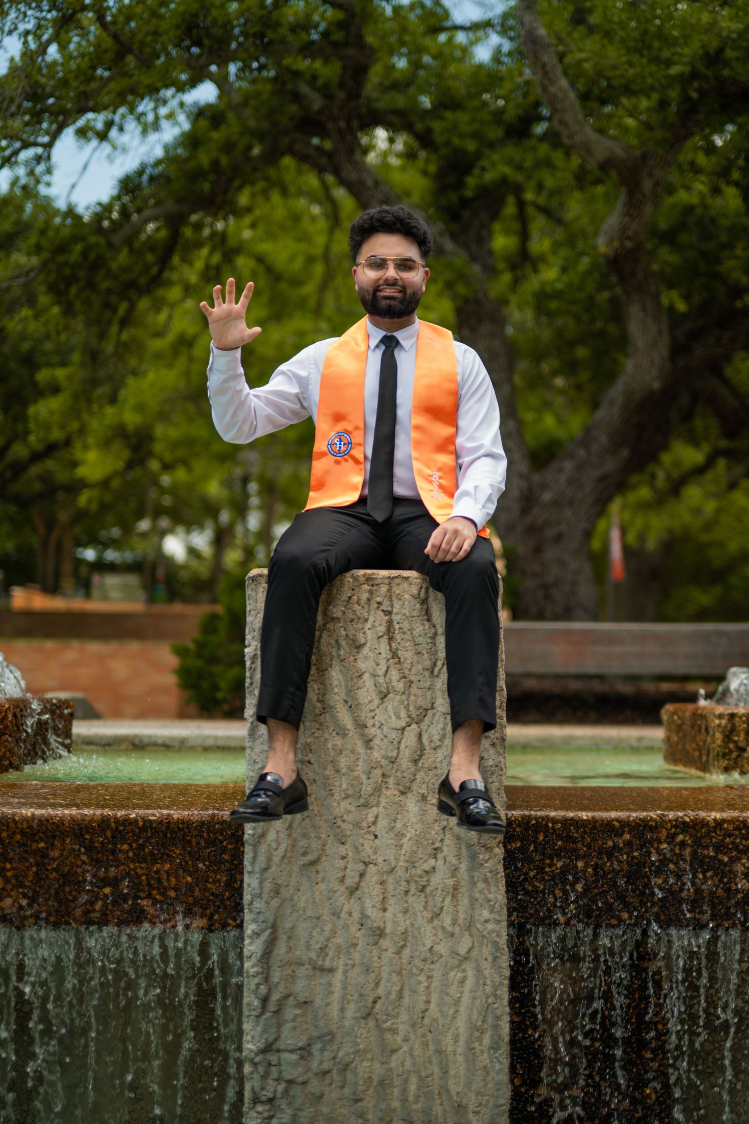 Graduate poses cheerfully on stone wall wearing orange stole and formal attire.