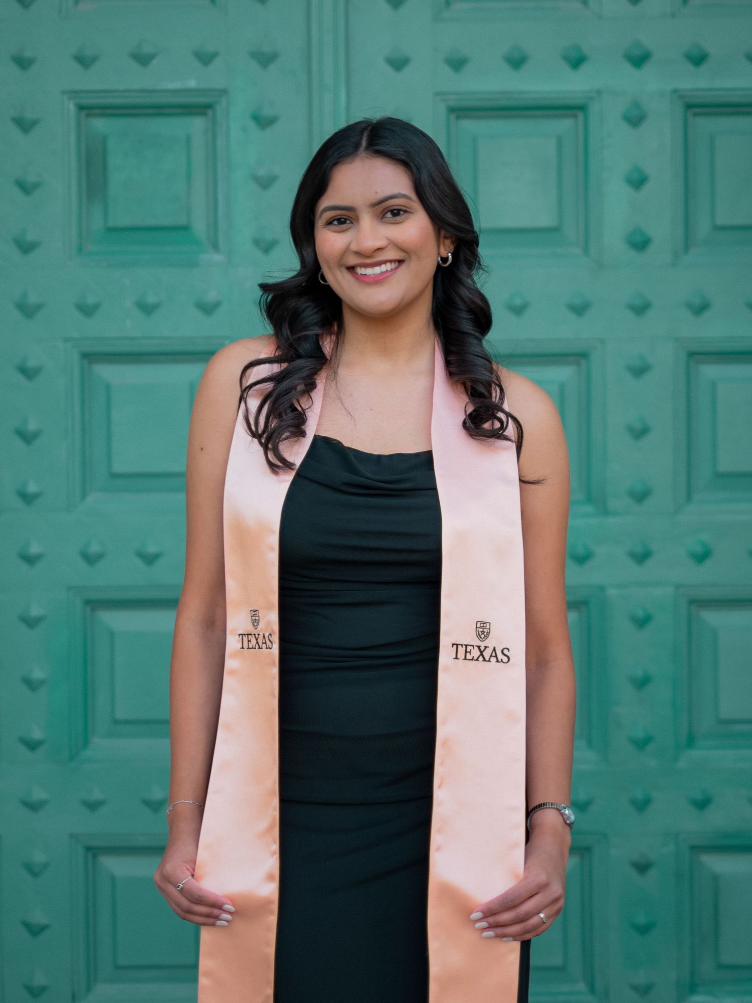 Graduate in black dress and pink sash poses in front of turquoise doors during graduation photo session.