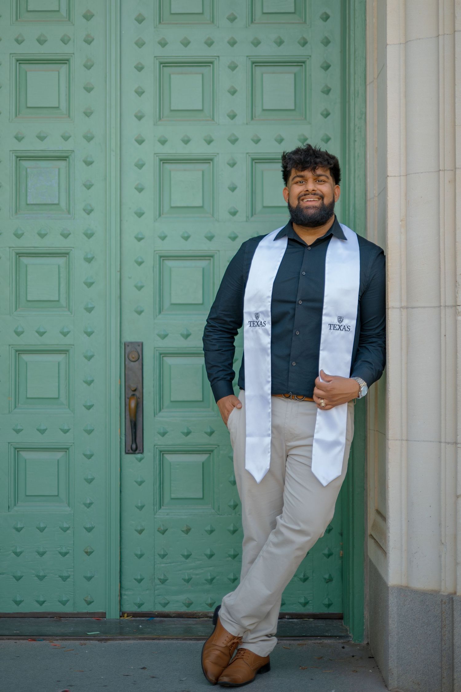 Graduate poses playfully against mint green ornate doors in graduation attire.