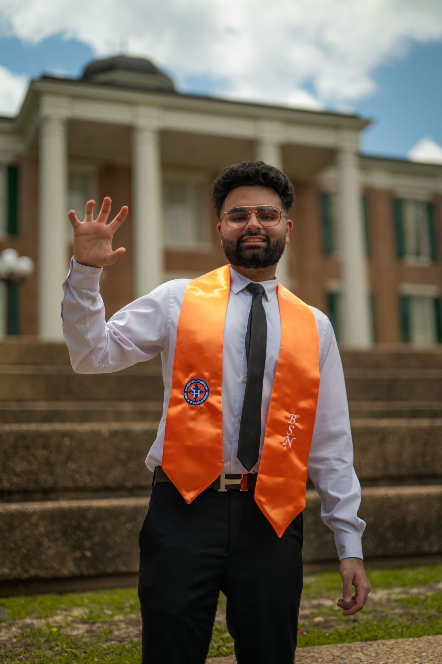 Graduate in formal attire wearing an orange graduation stole poses joyfully on campus grounds.