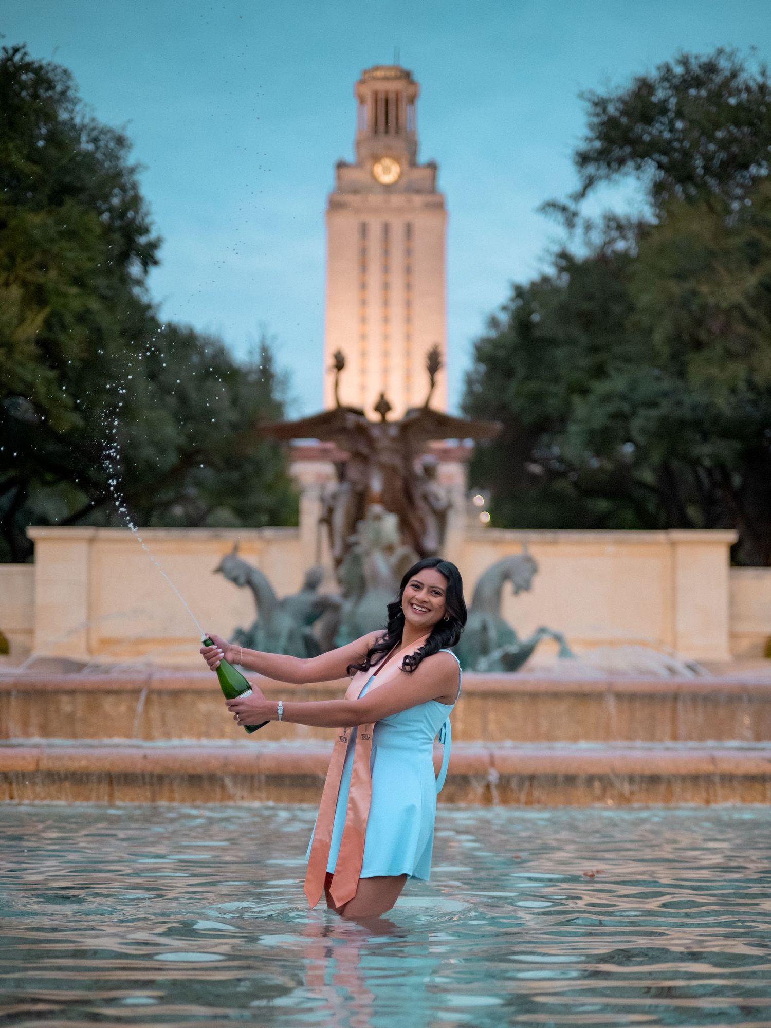 A person in a light blue dress relaxes in the water with the iconic UT Austin Tower illuminated in the background.