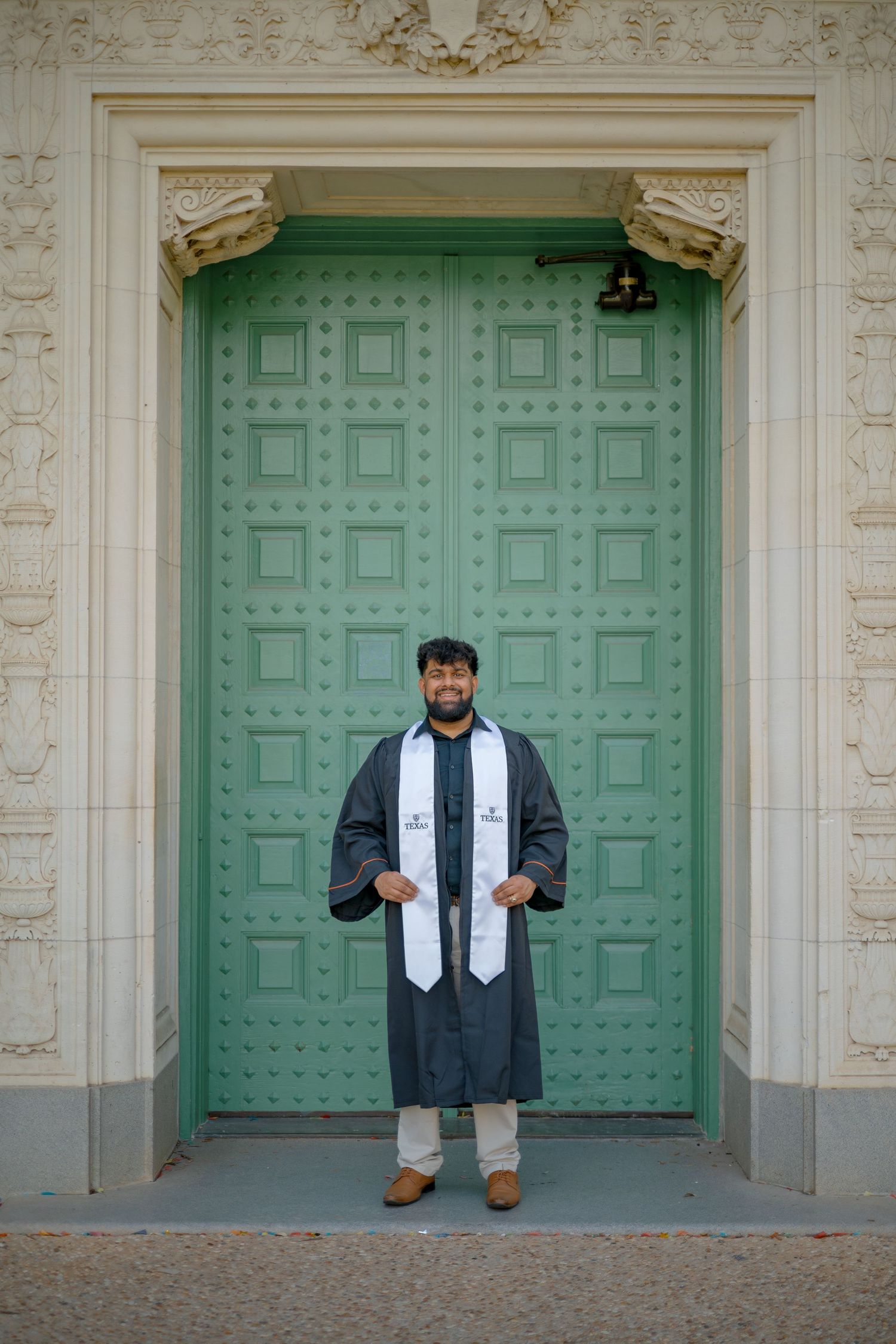 Graduate stands centered between ornate mint green doors in full graduation attire.
