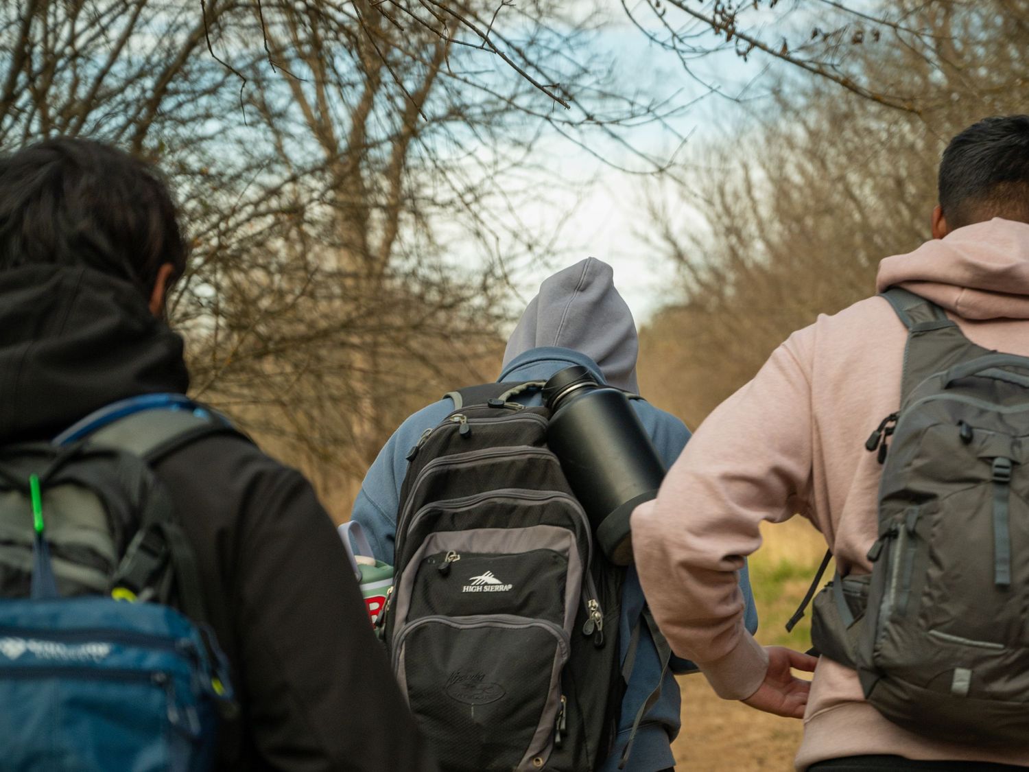 Row of backpack-wearing hikers viewed from behind on an outdoor trail.