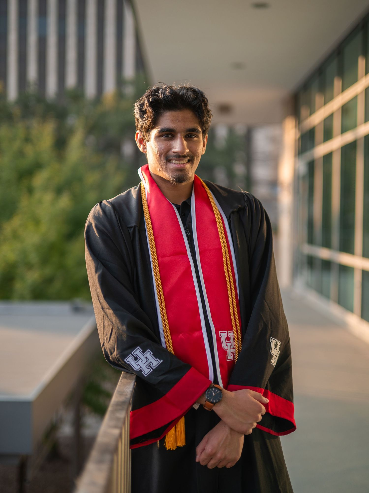 Graduate in black gown and red stole poses in outdoor hallway during golden hour.