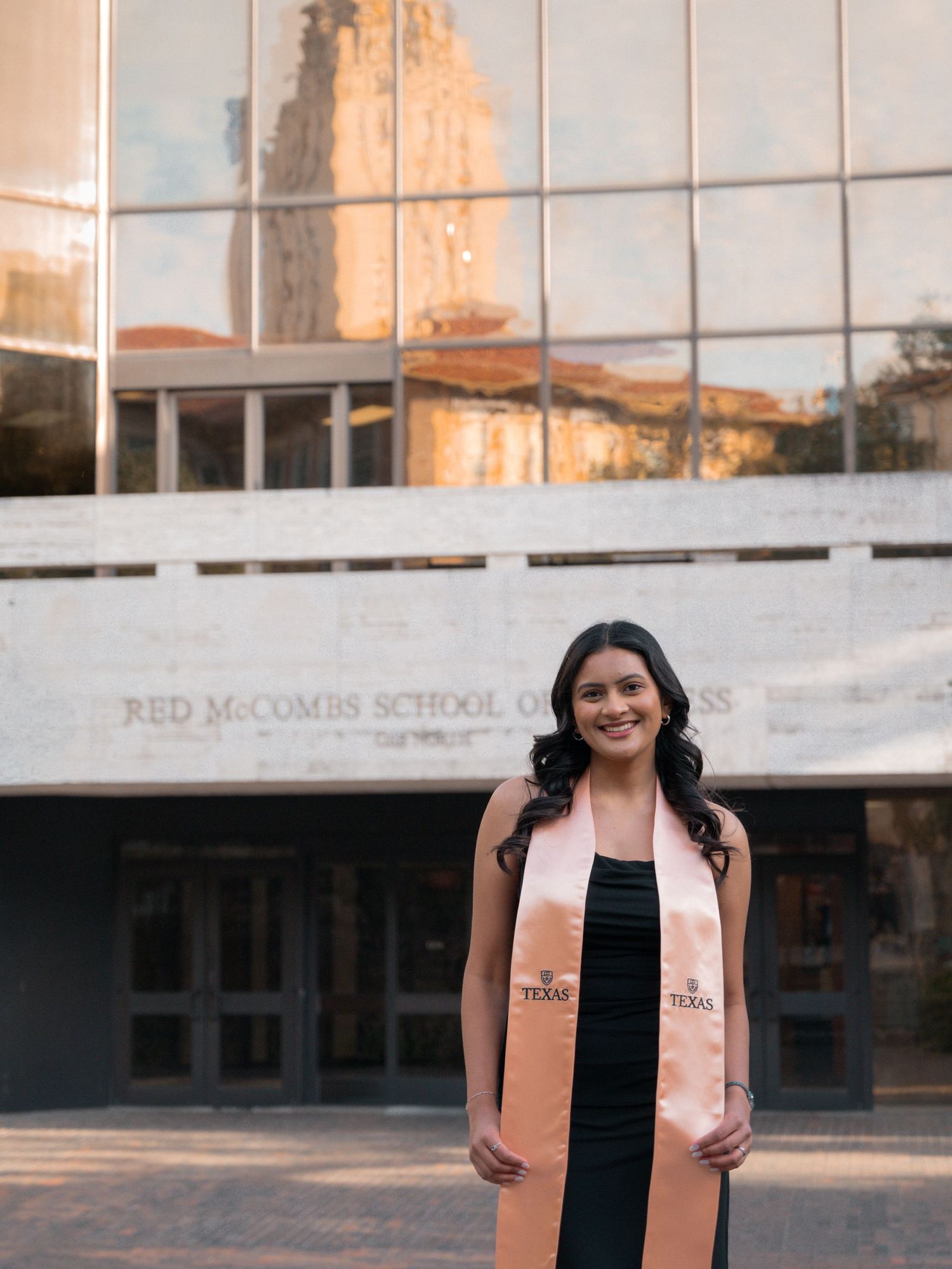 Graduate in a peach-colored stole stands smiling outside a modern university building at sunset.