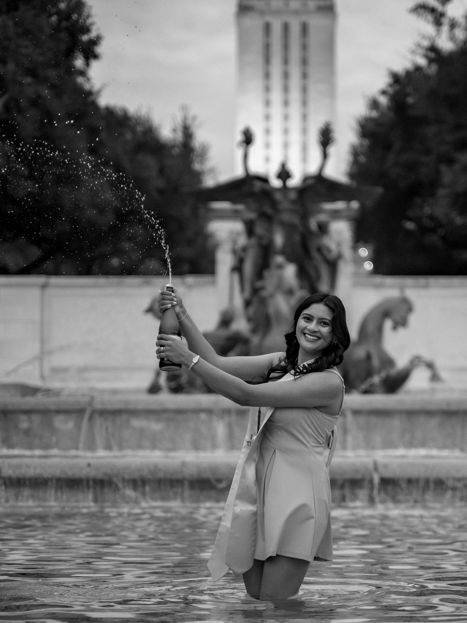 A person stands playfully in a fountain while spraying water, with a university tower and ornate lamp post visible in background.