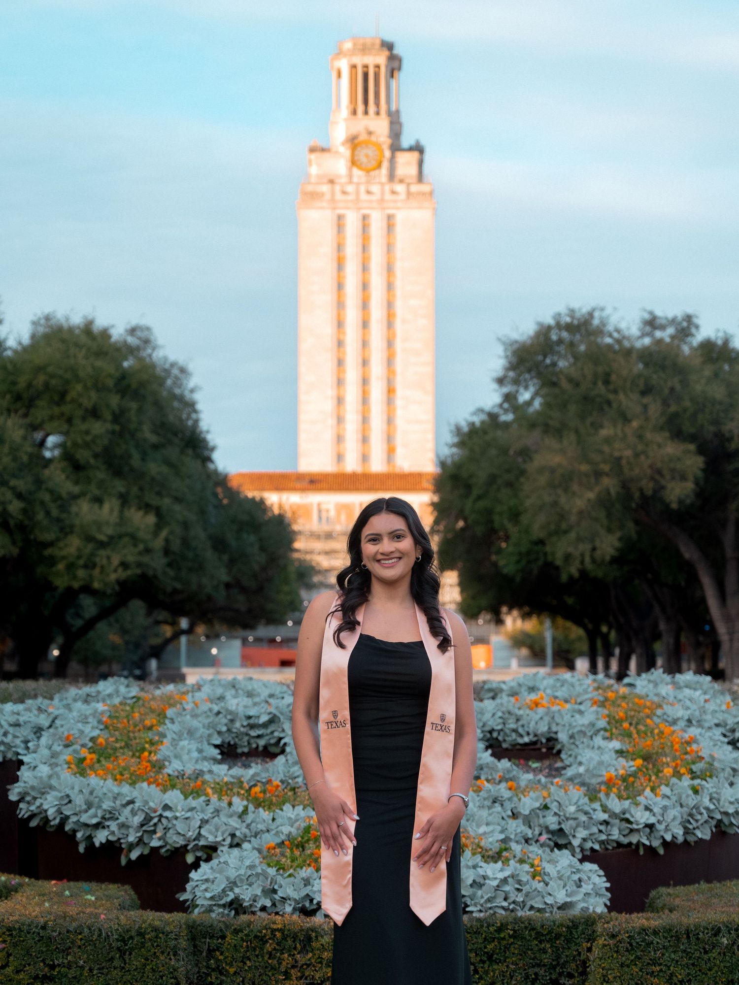 A graduation photo taken in front of the iconic University of Texas Tower with blue and gold flowers in the foreground.