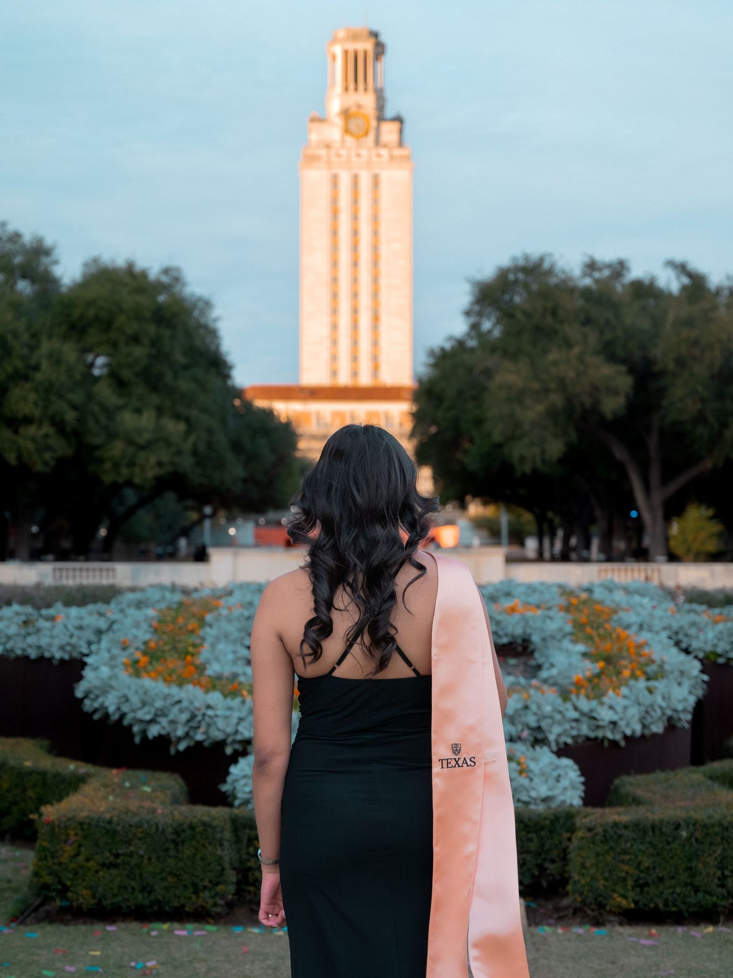 Iconic UT Austin Tower seen from campus with landscaped bluebonnets and trees in foreground during sunset.