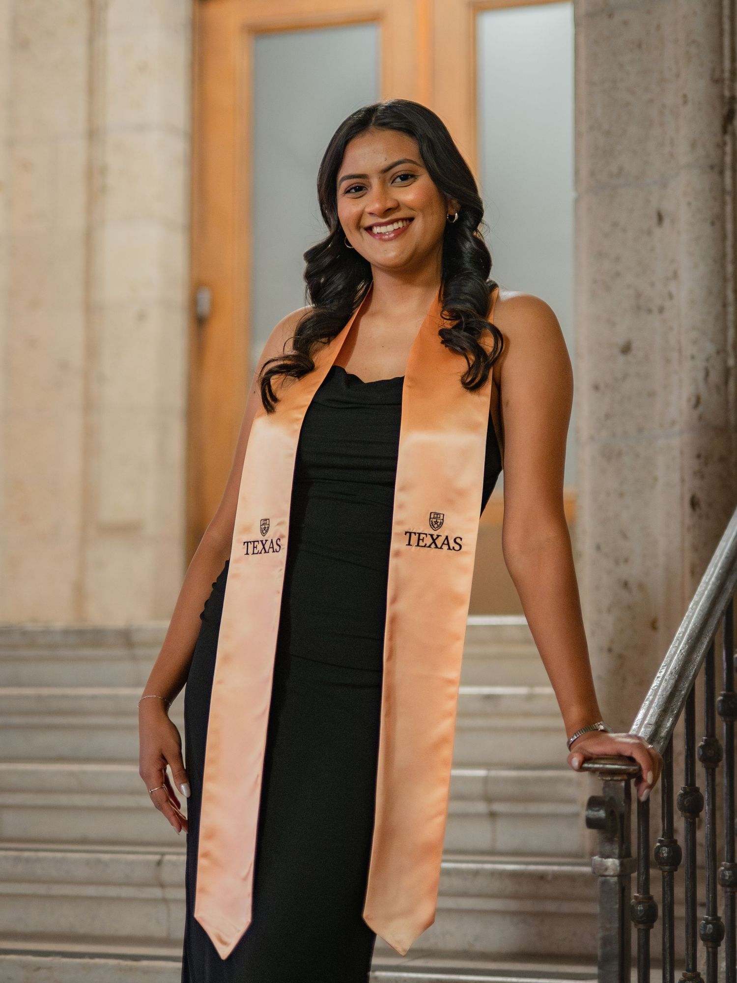 Graduate in black dress and peach-colored graduation stole poses for photos on stone steps.