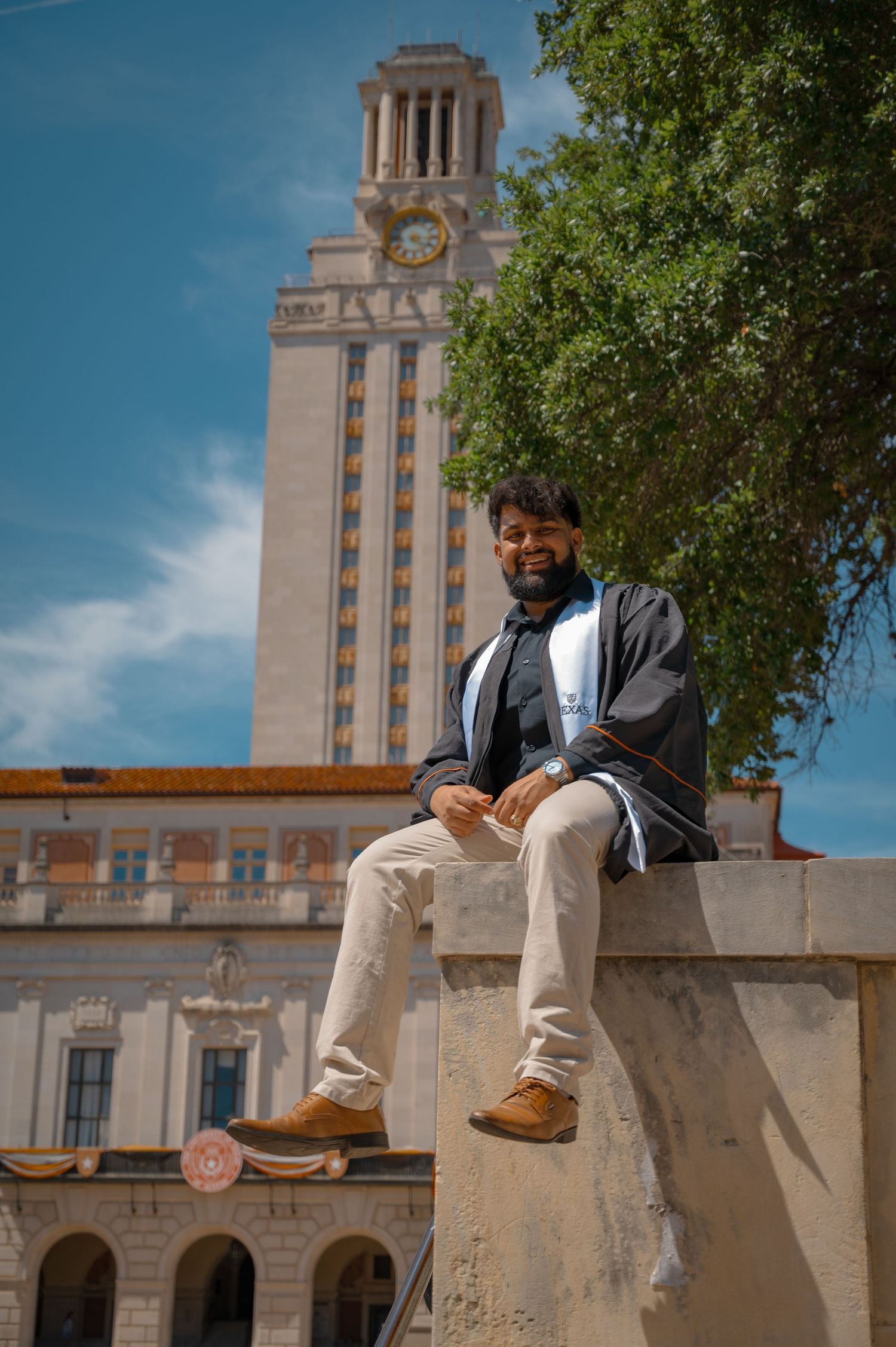 Graduate sits casually on stone ledge with University of Texas tower visible behind.