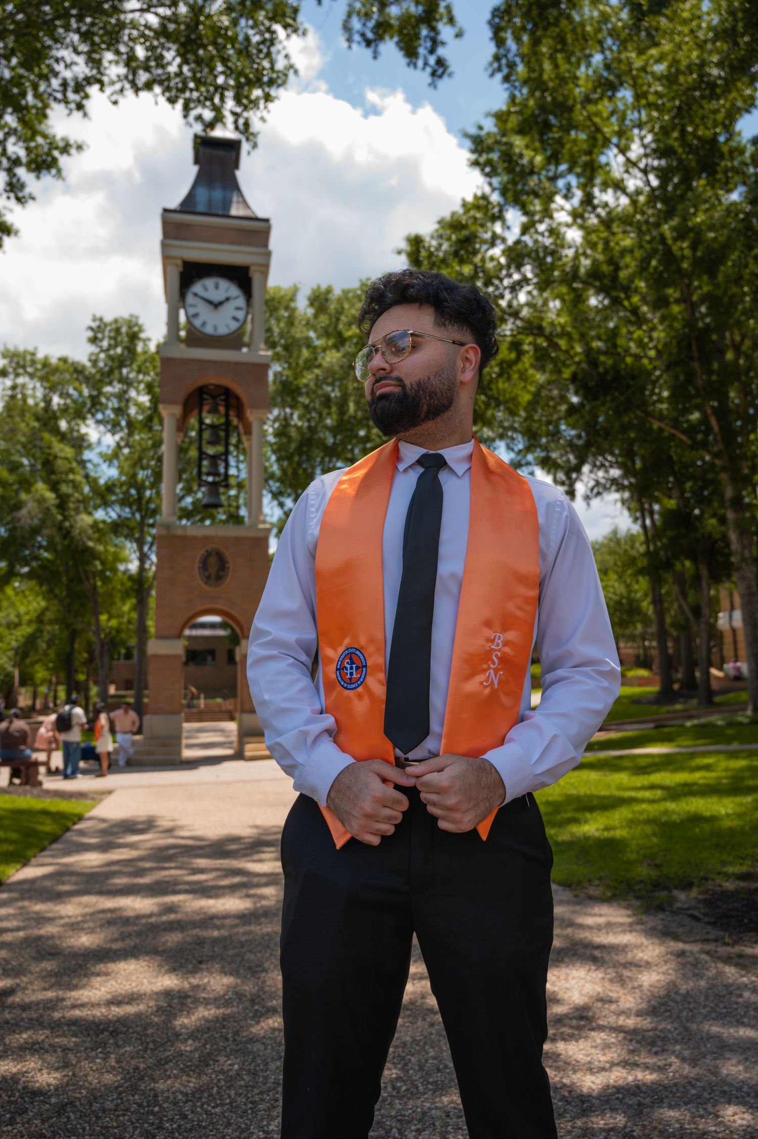Graduate in orange stole and formal wear poses on campus with clock tower in background.