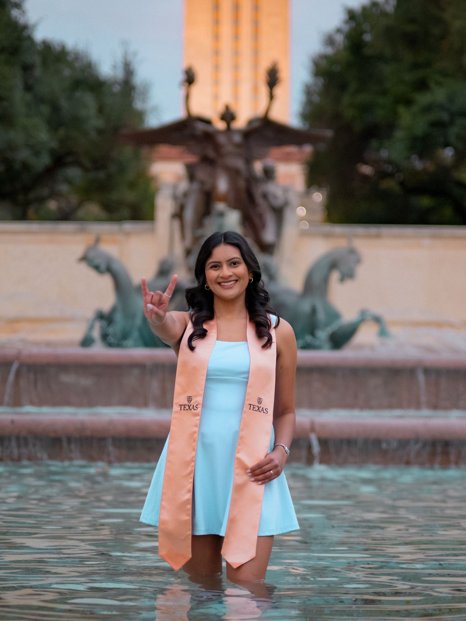 A person in a light blue dress and peach blazer poses in a decorative fountain with ornate sculptures in the background.