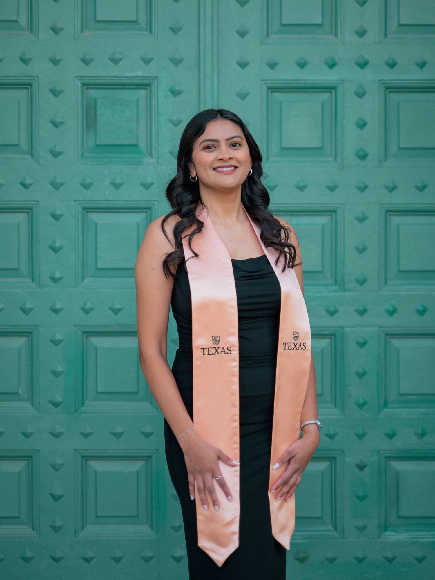 A graduate in black dress and peach stole poses against a decorative turquoise door background.
