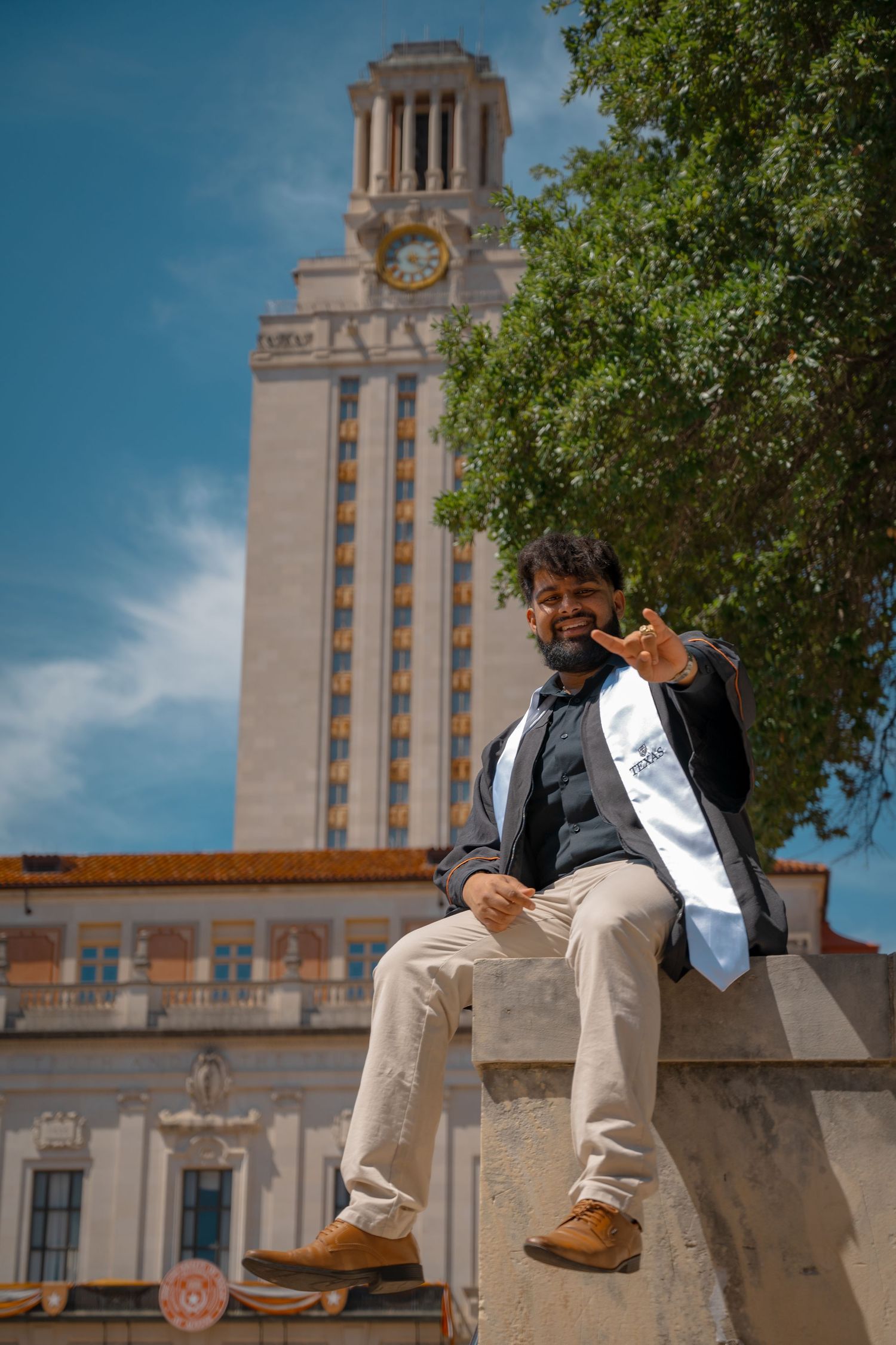 Graduate in black jacket and white sash poses on ledge with UT Austin Tower in background.