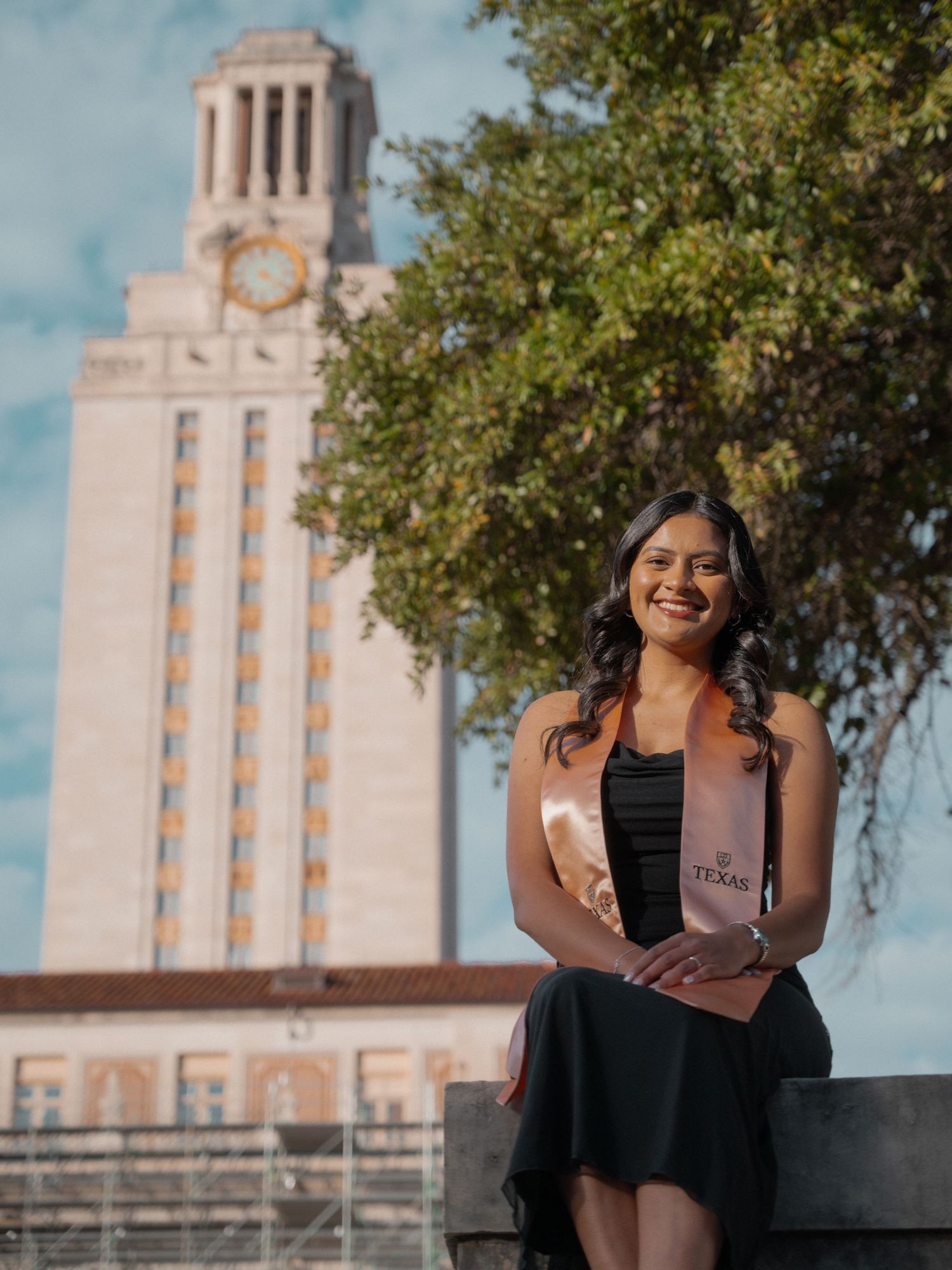 A person in a black top poses in front of the University of Texas Tower on a sunny day with trees in view.