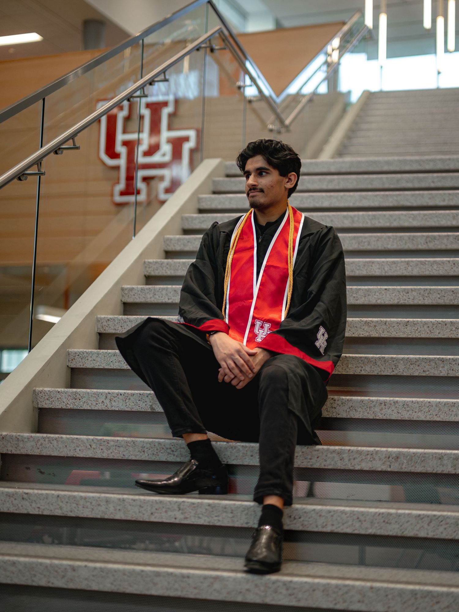 Graduate in black cap and gown with red stole sits contemplatively on concrete steps.