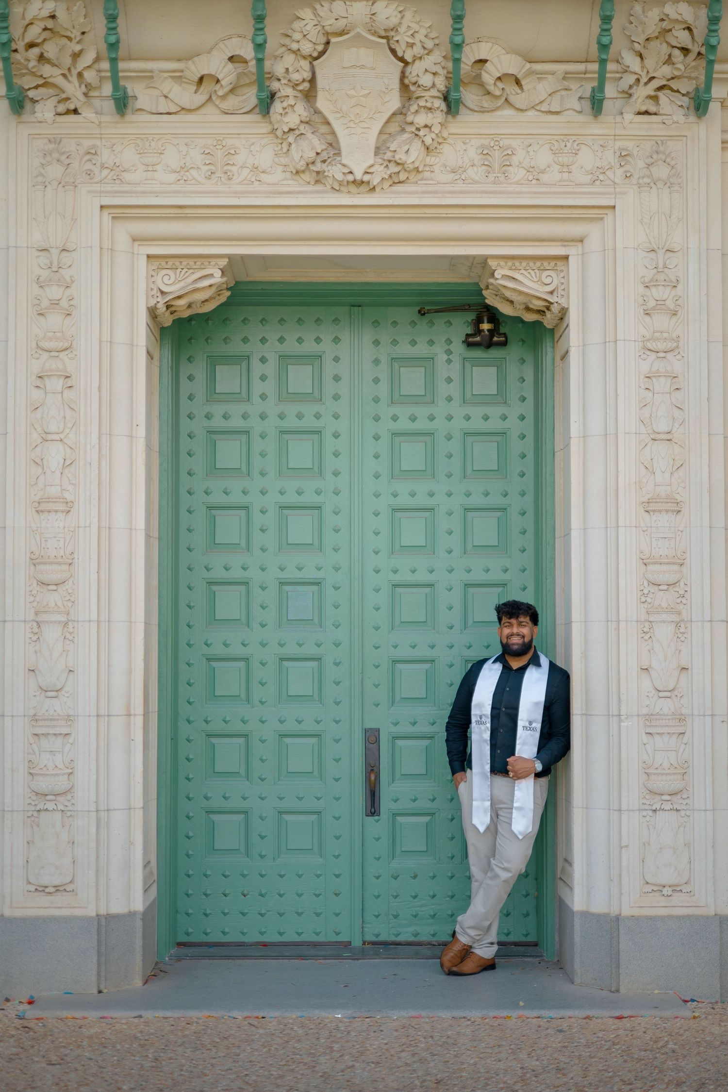 Wide shot of graduate posing by decorative mint green doors with classical architectural details.