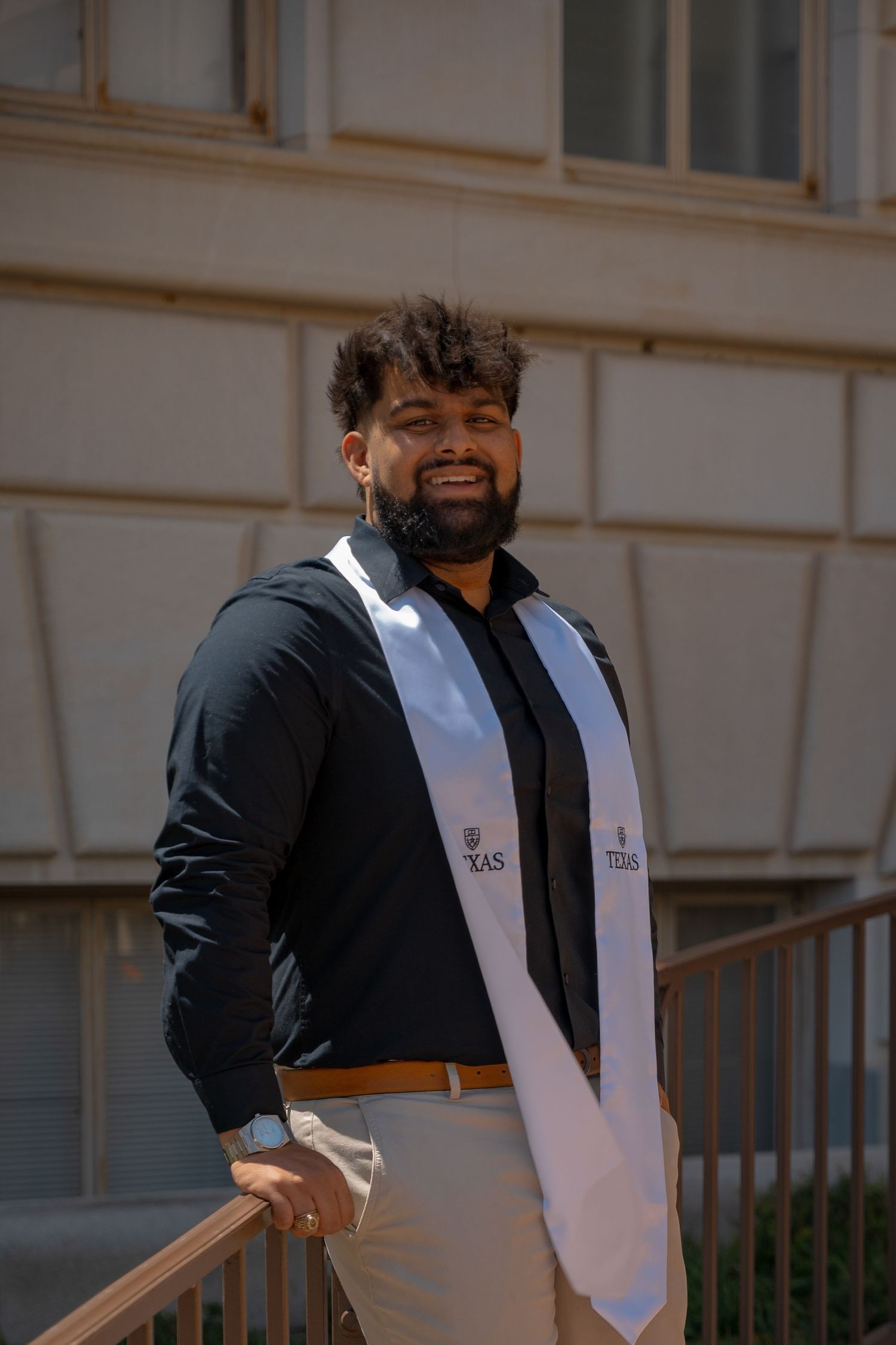 Graduate in black jacket with Texas sash poses on campus steps.