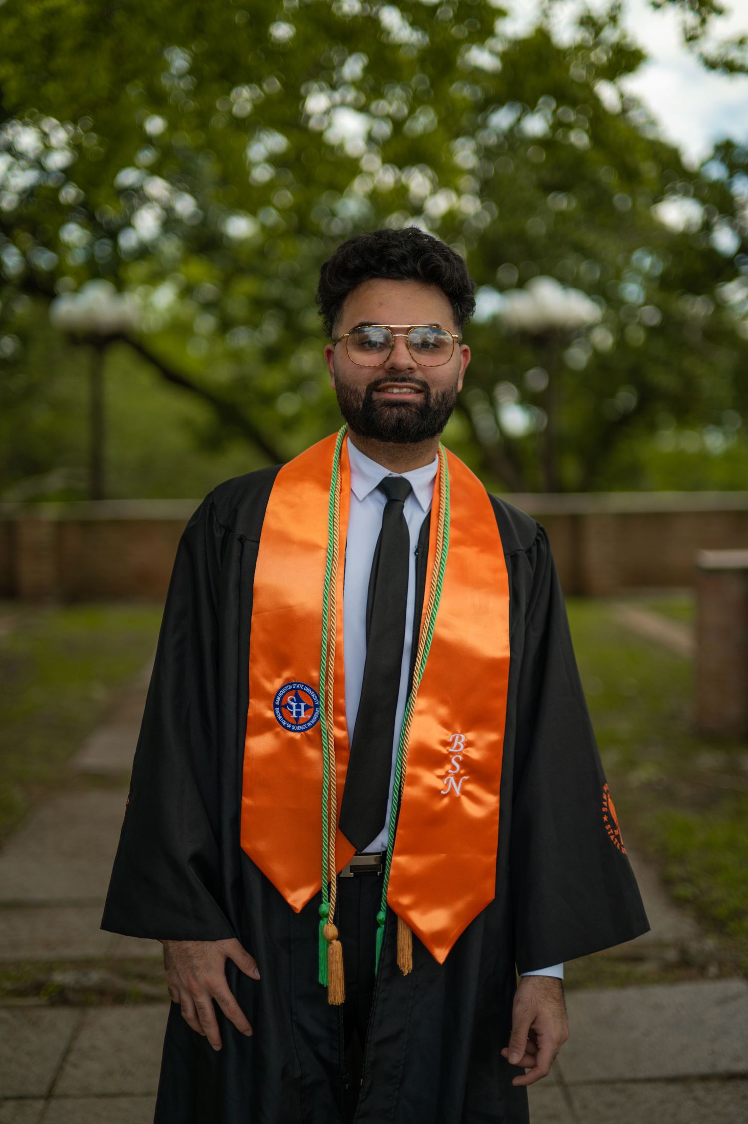 Graduate wearing academic regalia with orange stole and glasses poses outdoors on campus.